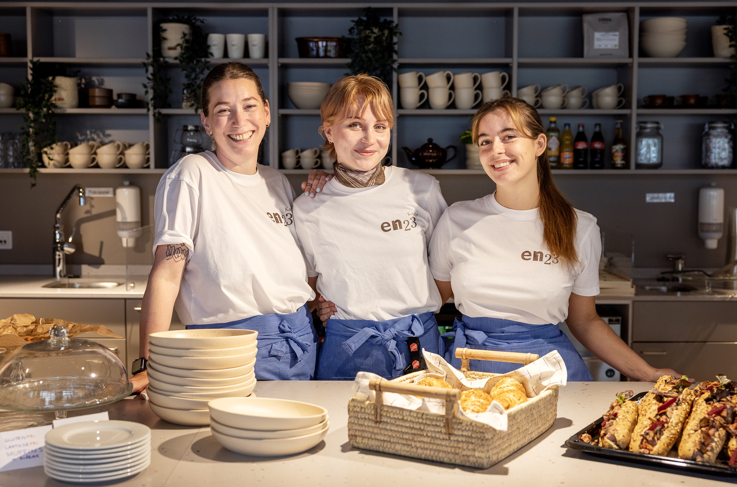 Foto: Tre kvinner i hvite T-skjorter med logoen til Kafé En23 står bak en kafédisk. Alle har på seg blå forklær. I bakgrunnen sees hyller med kopper og glass.