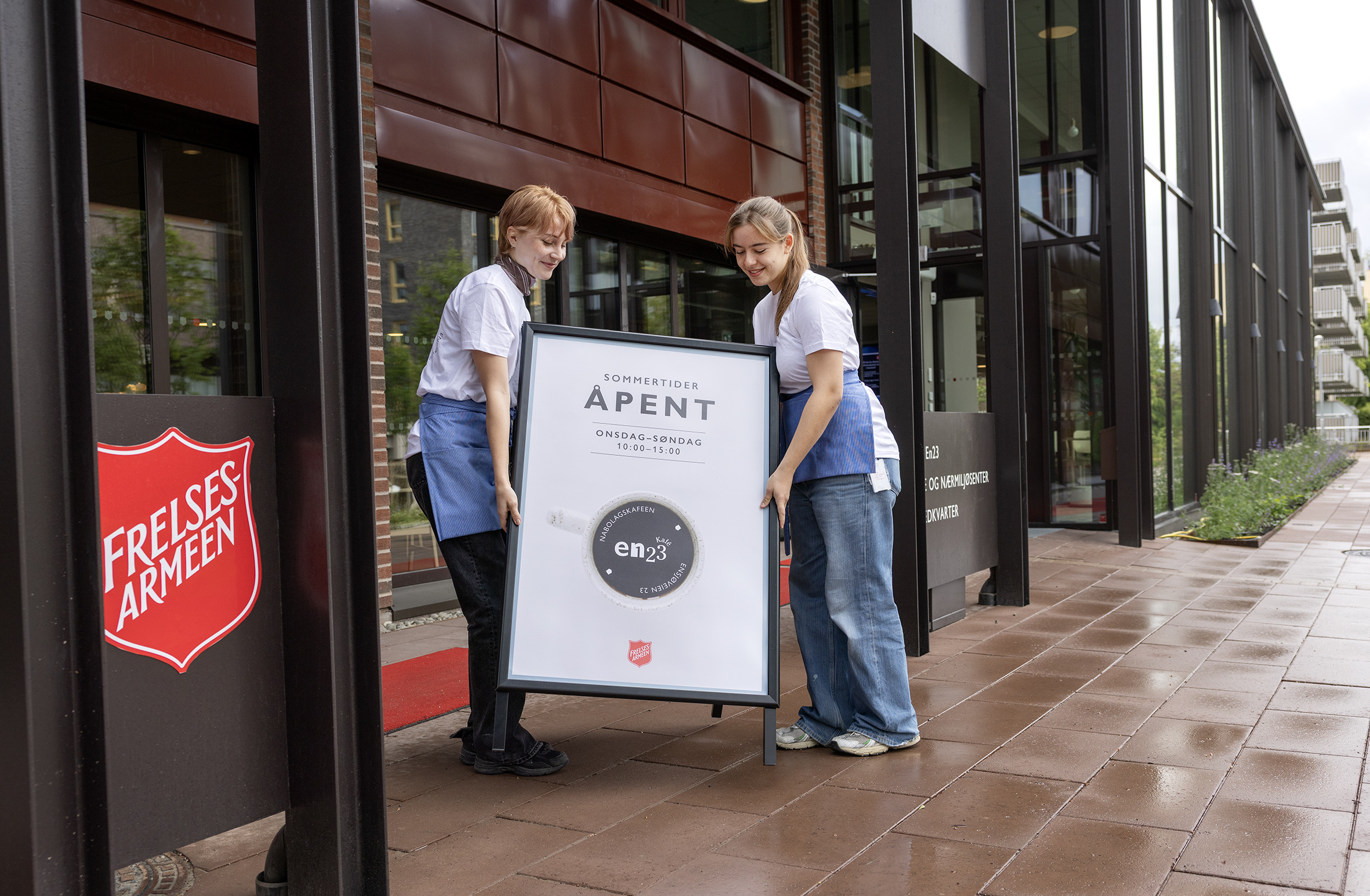 Foto: To unge kvinner bærer et skilt ut med logoen til kafeen. Like ved siden av er logoen til Frelsesarmeen også.