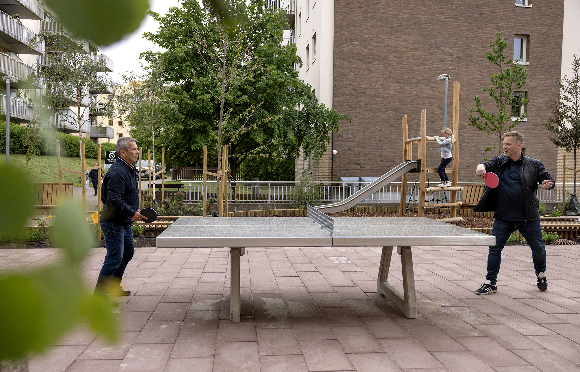Foto: To voksne menn som spiller bordtennis ute på et beplantet torg.