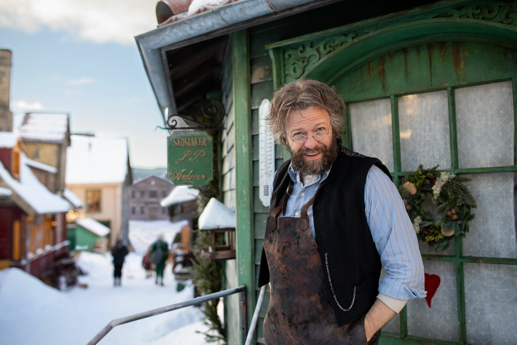 Middelaldrende mann med briller og skjegg står på trappa til et gammelt, julepyntet hus. Ute er det snø i gata.
