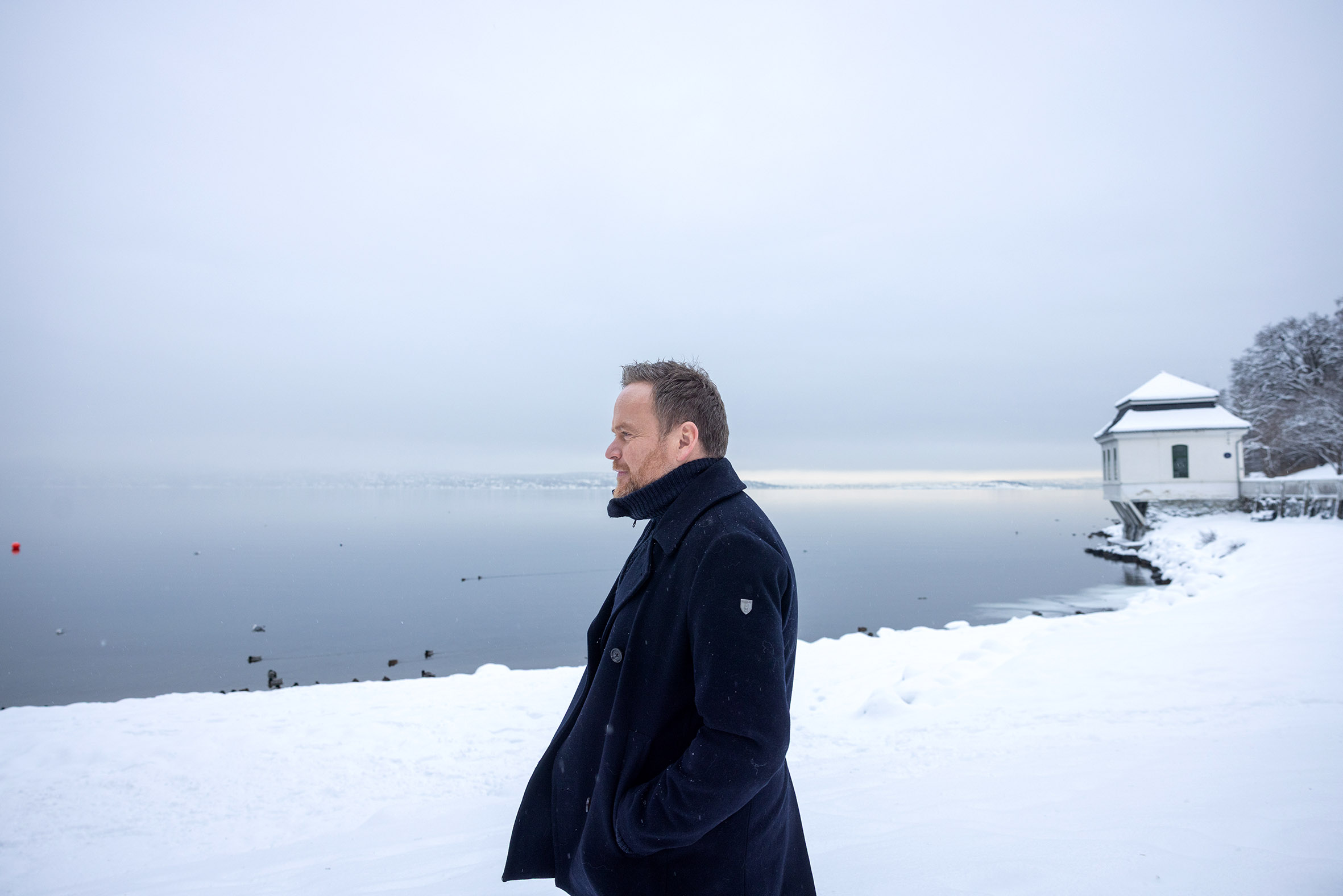 Foto: Mann i blå vinterjakke går bortover en flat mark med en snødekket strand i bakgrunnen og et hvitt, gammelt badehus i bakgrunnen.