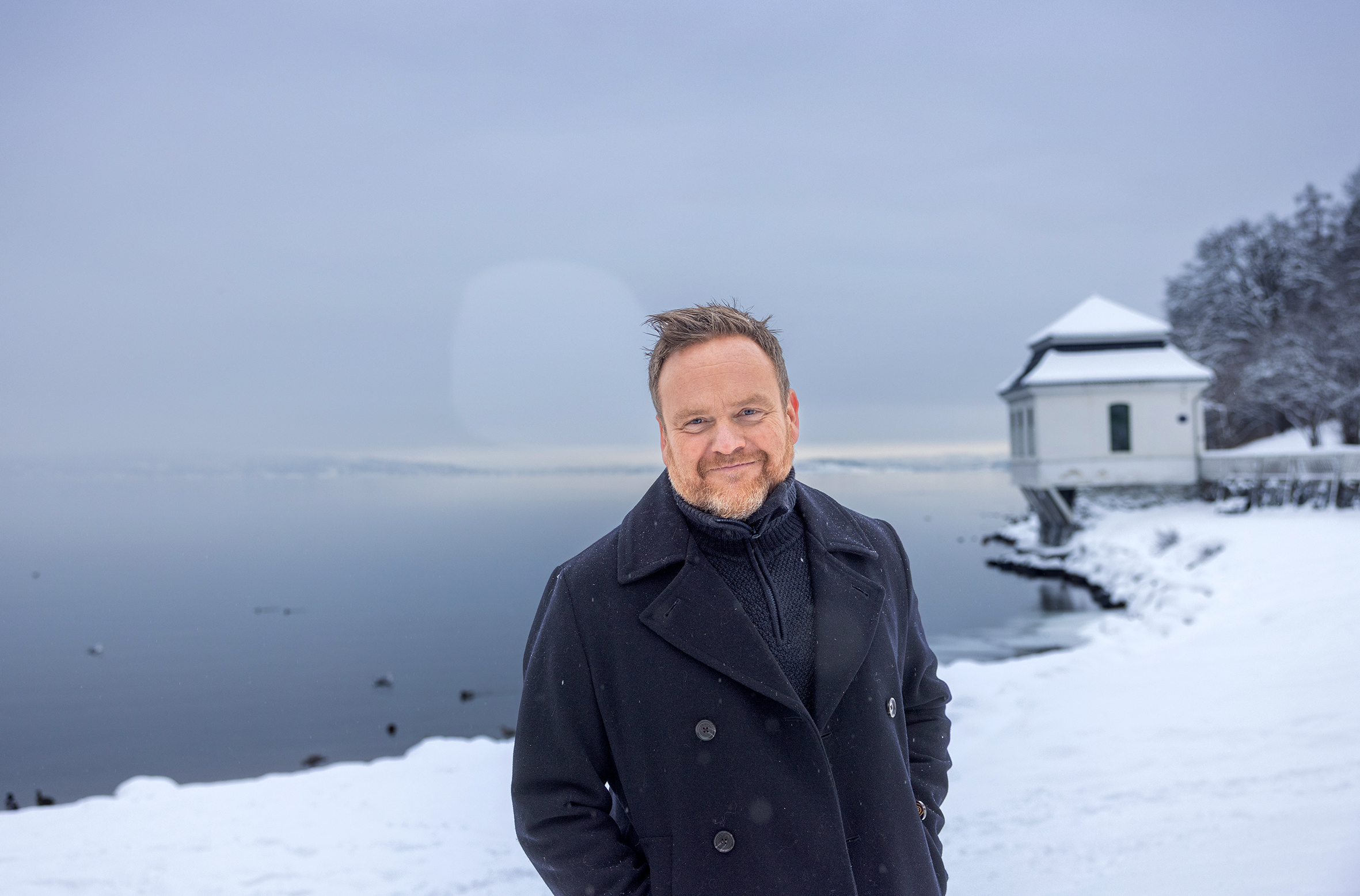 Foto: Mann i åpen, blå jakke står i snøen med snødekket strand og en gammelt, hvitt badehus bak.