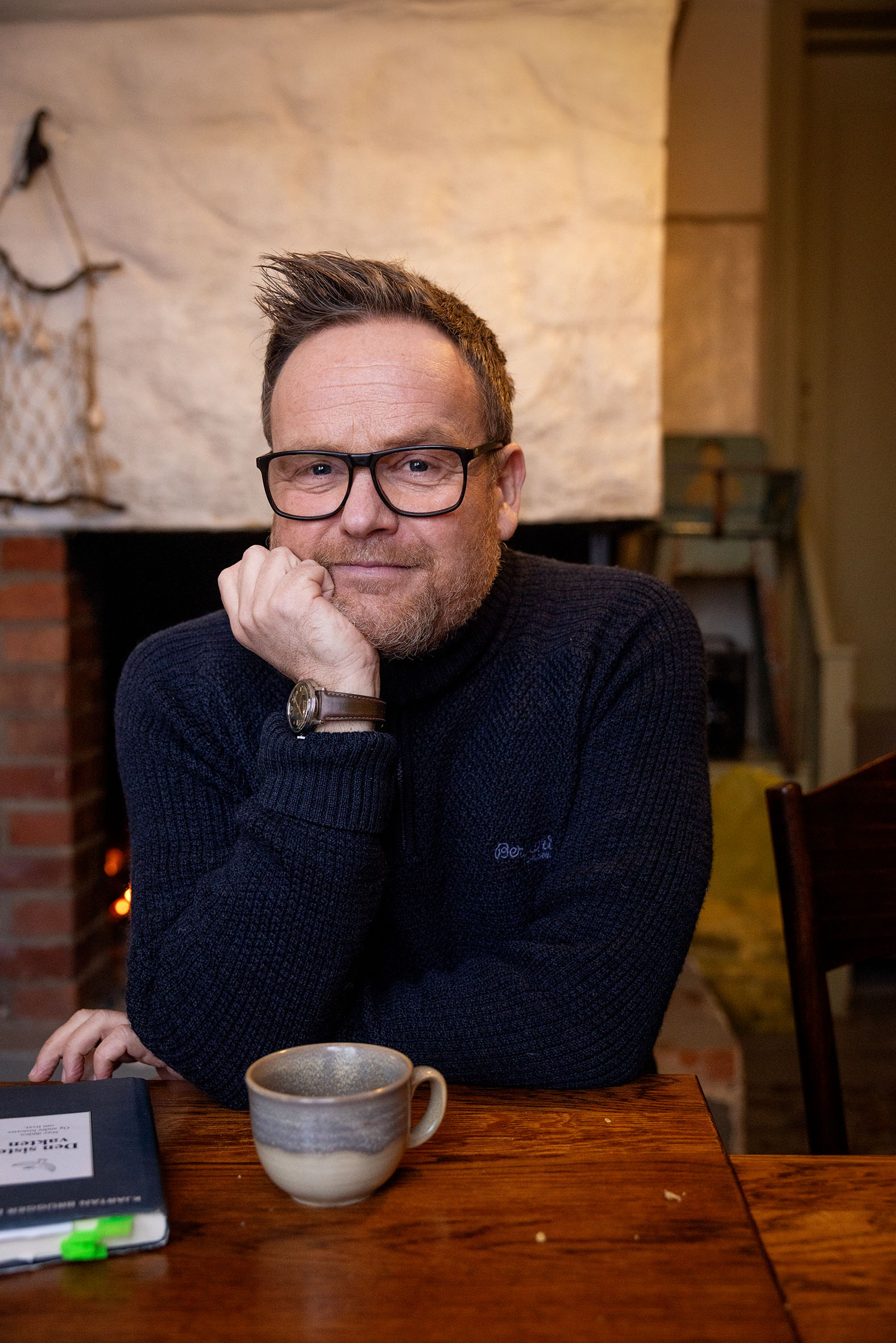 Foto: Mann med briller og blå genser sitter ved et trebord. Bak ham er det en peis.