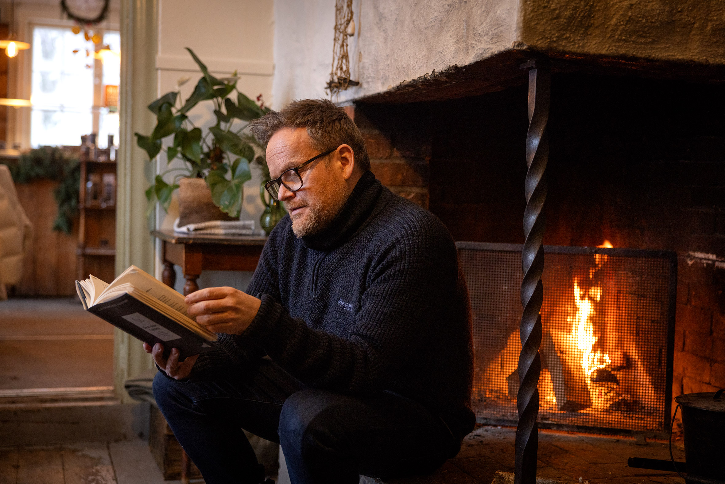 Foto: Mann sitter på kanten av en gammel, oppmurt peis med flammer i. Lokalene er gamle og i tre. I enden av det lille lokalet står en kjøpmannsdisk med kaker på.