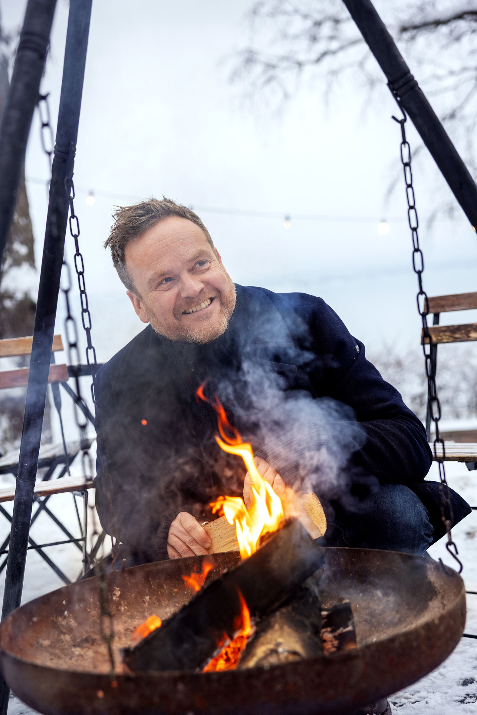 Foto: Mann med skjegg sitter på huk bak en bålpanne med fyr i ute. Det er snø på markene.