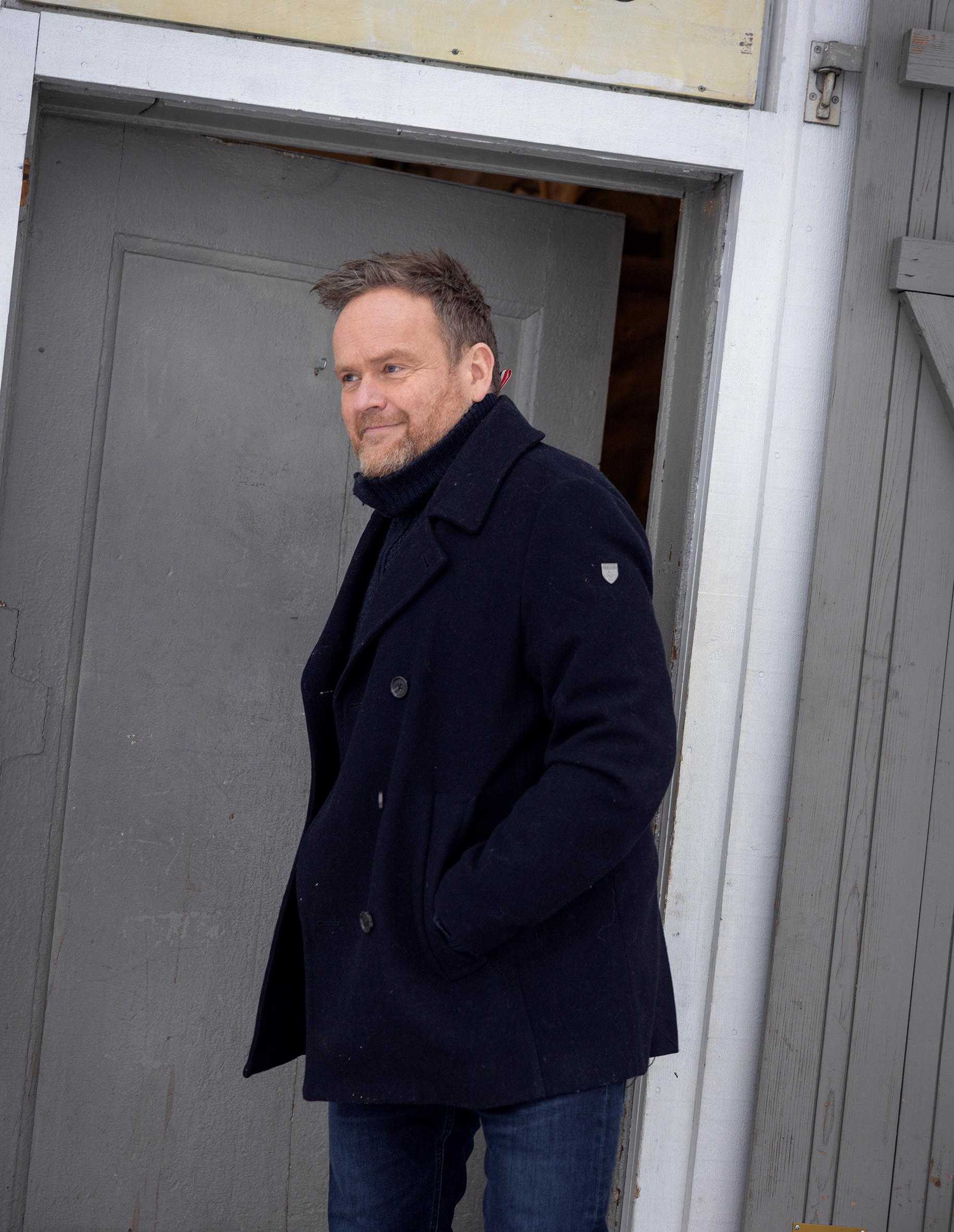 Foto: Blond mann med rødlig skjegg i 50-årene går ut av en grå dør. Han har på seg en marineblå vinterjakke i ull.