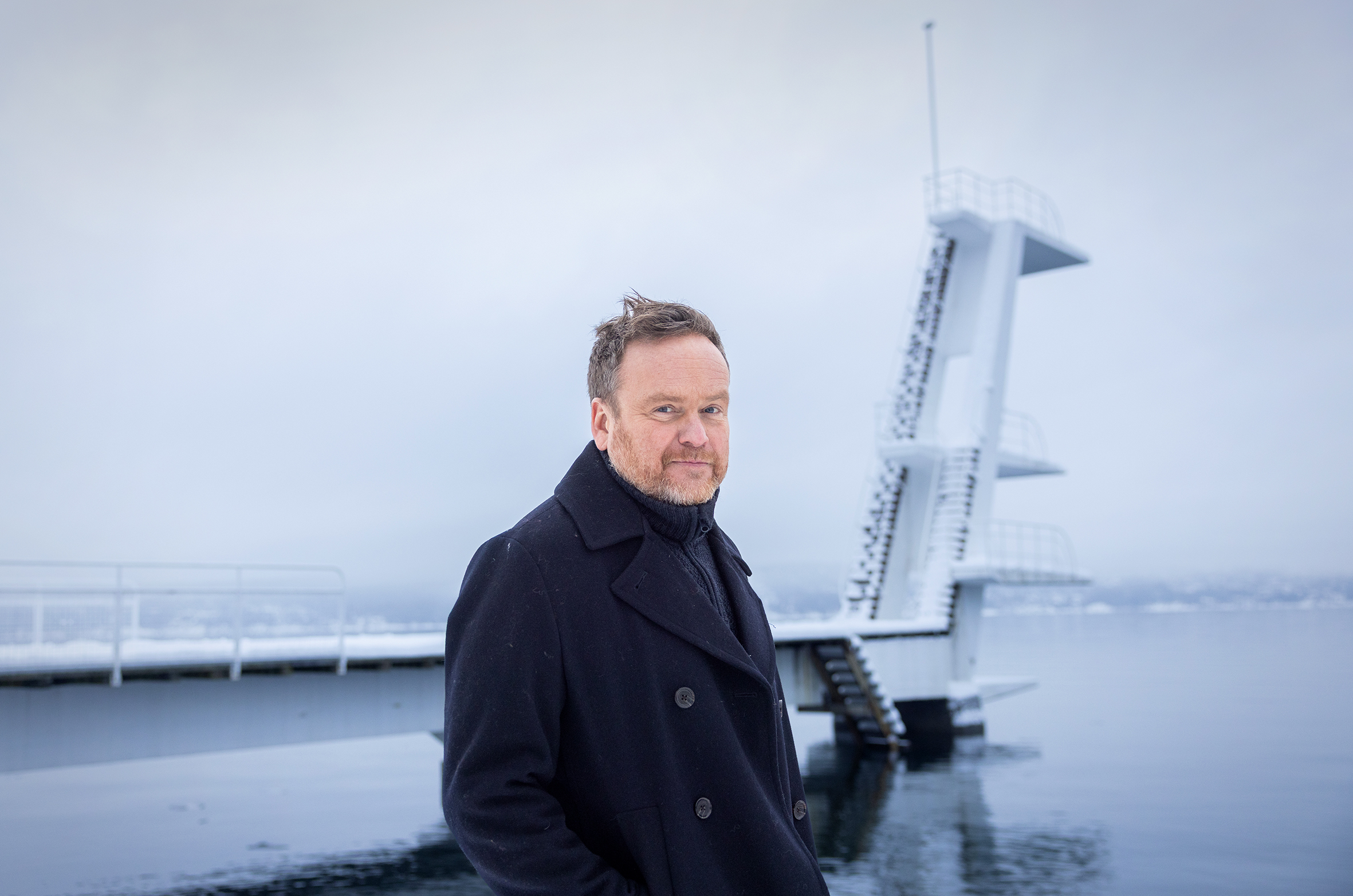 Foto: Vinterlandskap og et høyt stupetårn ytterst på en pir dekket av snø. I forkanten står en mann i 50-årene med blondt hår og rødlig skjegg i en blå vinterjakke. Han smiler litt lurt.