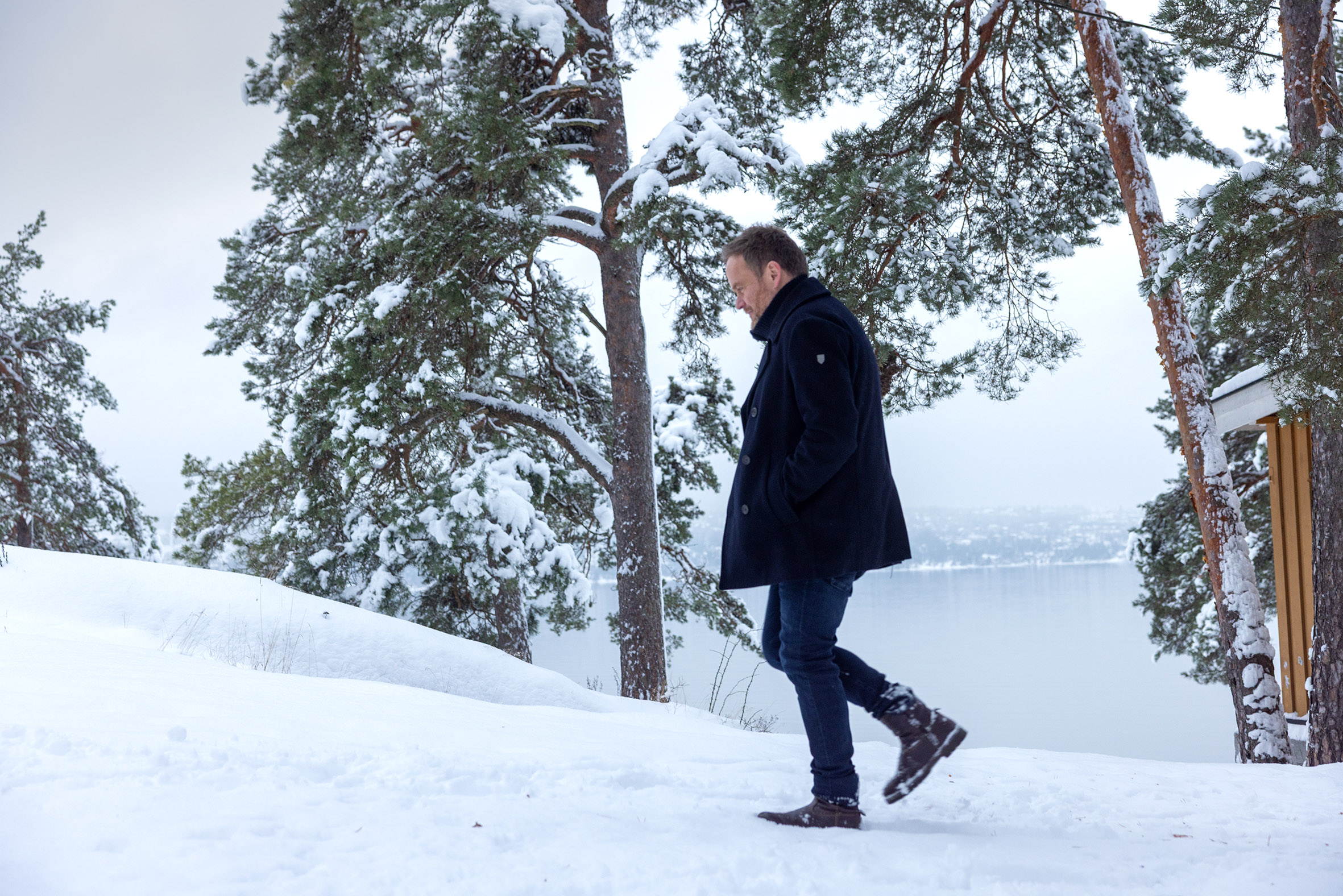 Foto: Mann med mørkeblå vinterjakke går oppover en snødekket bakke med litt av en gul vegg, furutrær og havet i bakgrunnen.