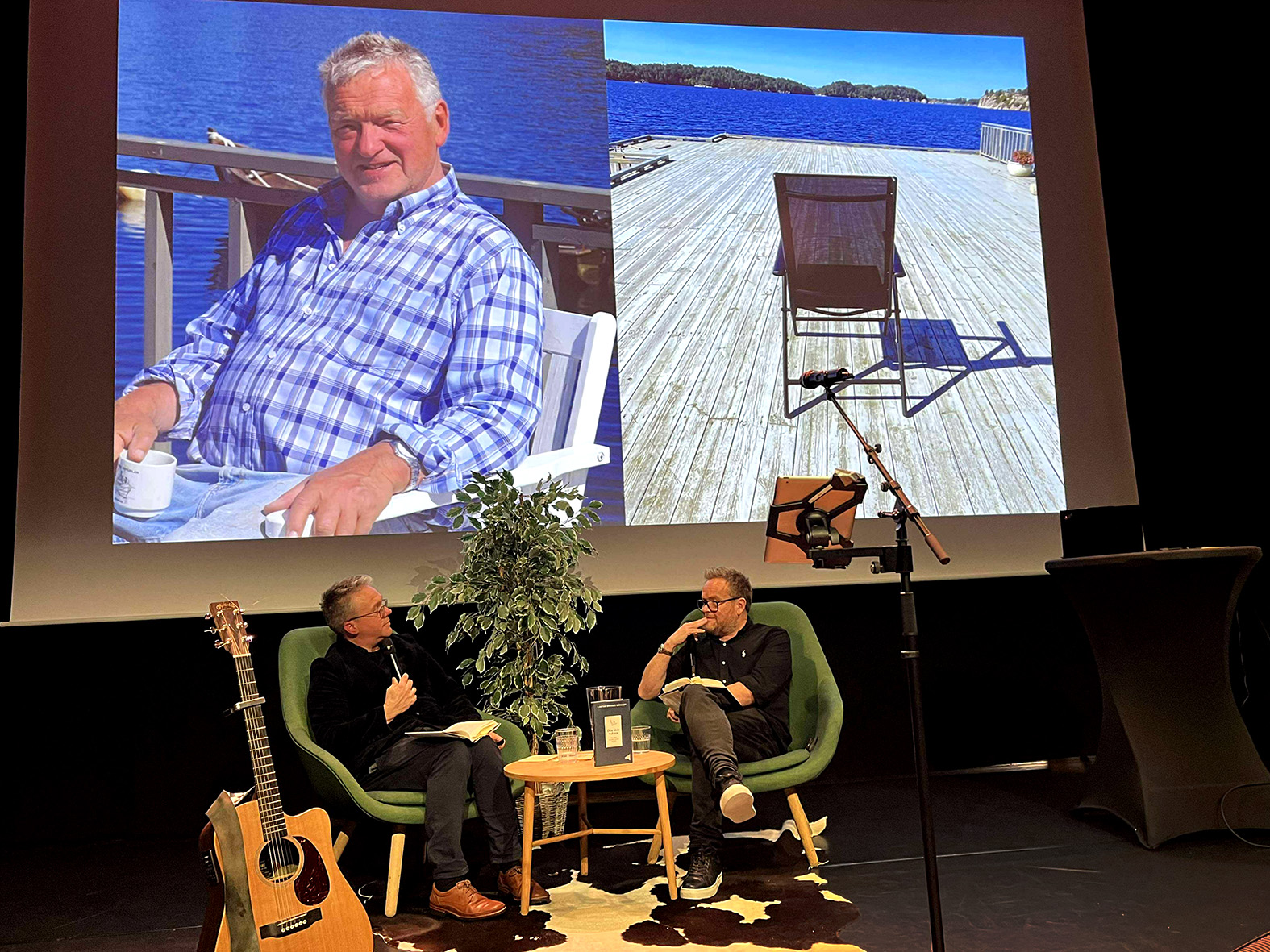 Foto: To menn med briller sitter i hver sin stol på en scene. På storskjermen bak er det bilde av en eldre, brunbarket mann i rutete skjorte og ved siden av et bilde av en tom på en terrasse i tre som strekker seg ut i havet.