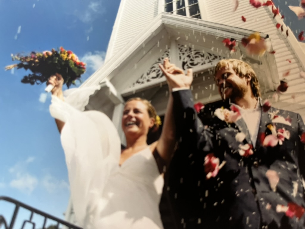 Foto: Brudepar tidlig i 30-årene en sommerdag med blå himmel. De står på trappa til en gammel, hvit kirke i tre og folk kaster ris og blomster på dem.