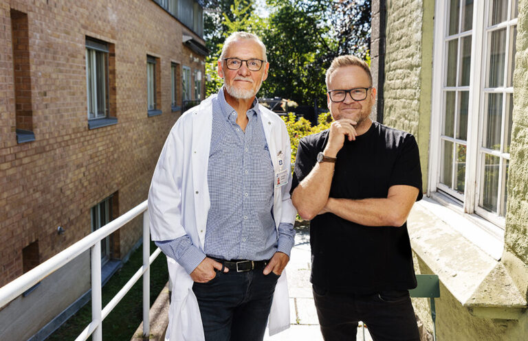 Foto: To menn med briller står ved siden av hverandre. Den ene er i 60-årene og den andre i 50-årene. Ene i hvit legefrakk oppå lyseblå skjorte og den andre i svart T-skjorte. De står ute på en gangbru mellom to gamle bygg i murstein.