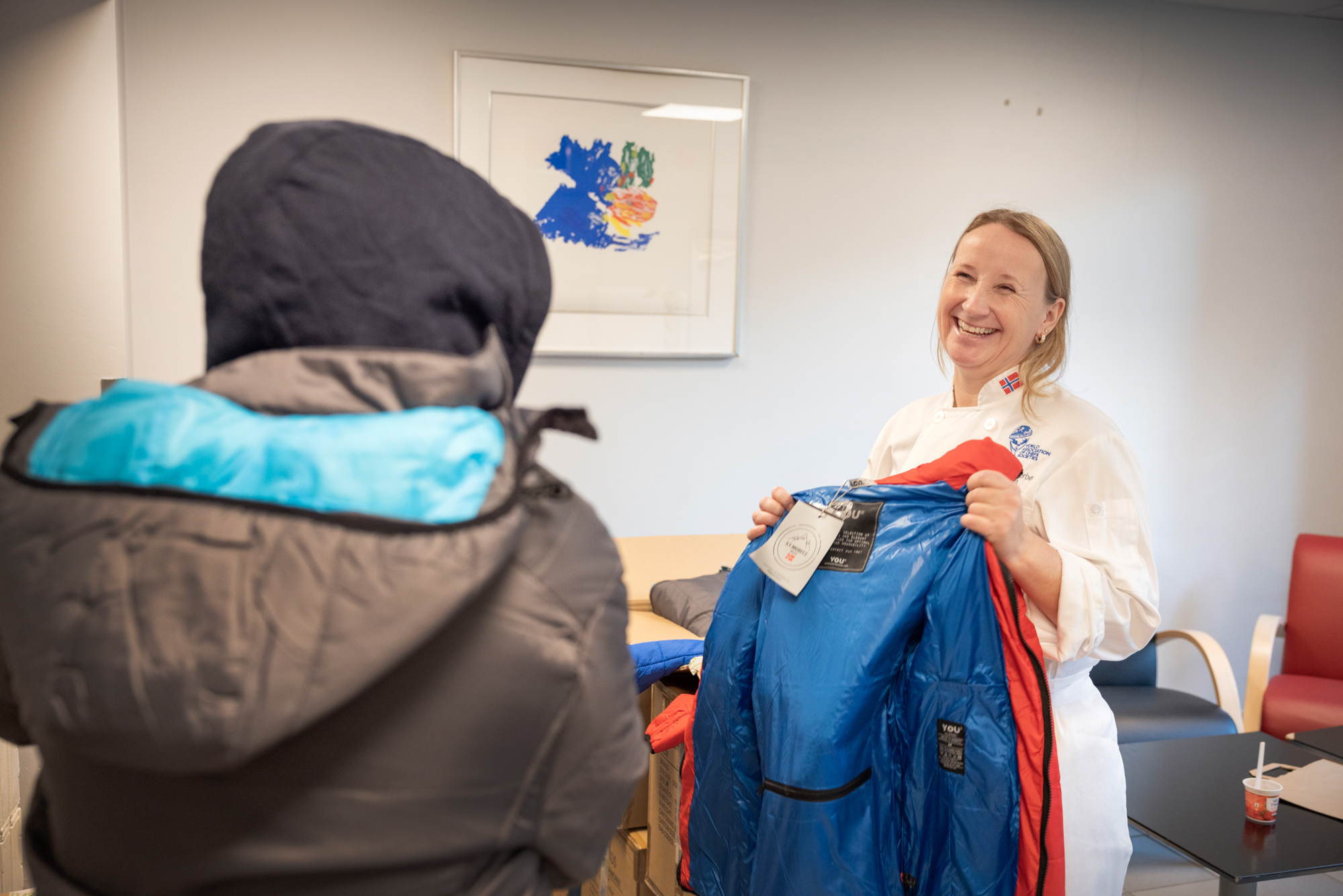 Kvinne i kokkeuniform holder opp en fargerik boblejakke. Vi ser rykken til en mann med hettegenser som prøver en jakke på.