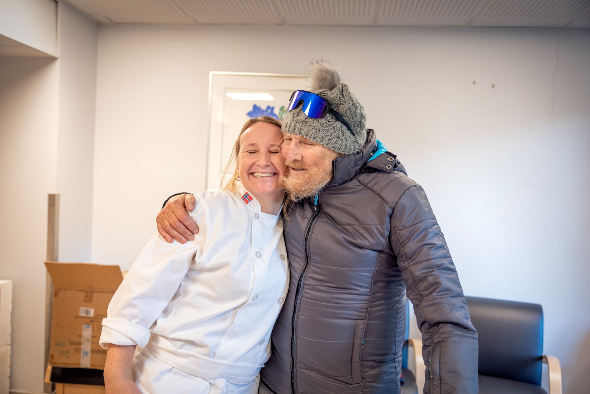 Mann i ny boblejakke klemmer den kvinnelige kokken. Kinn mot kinn.