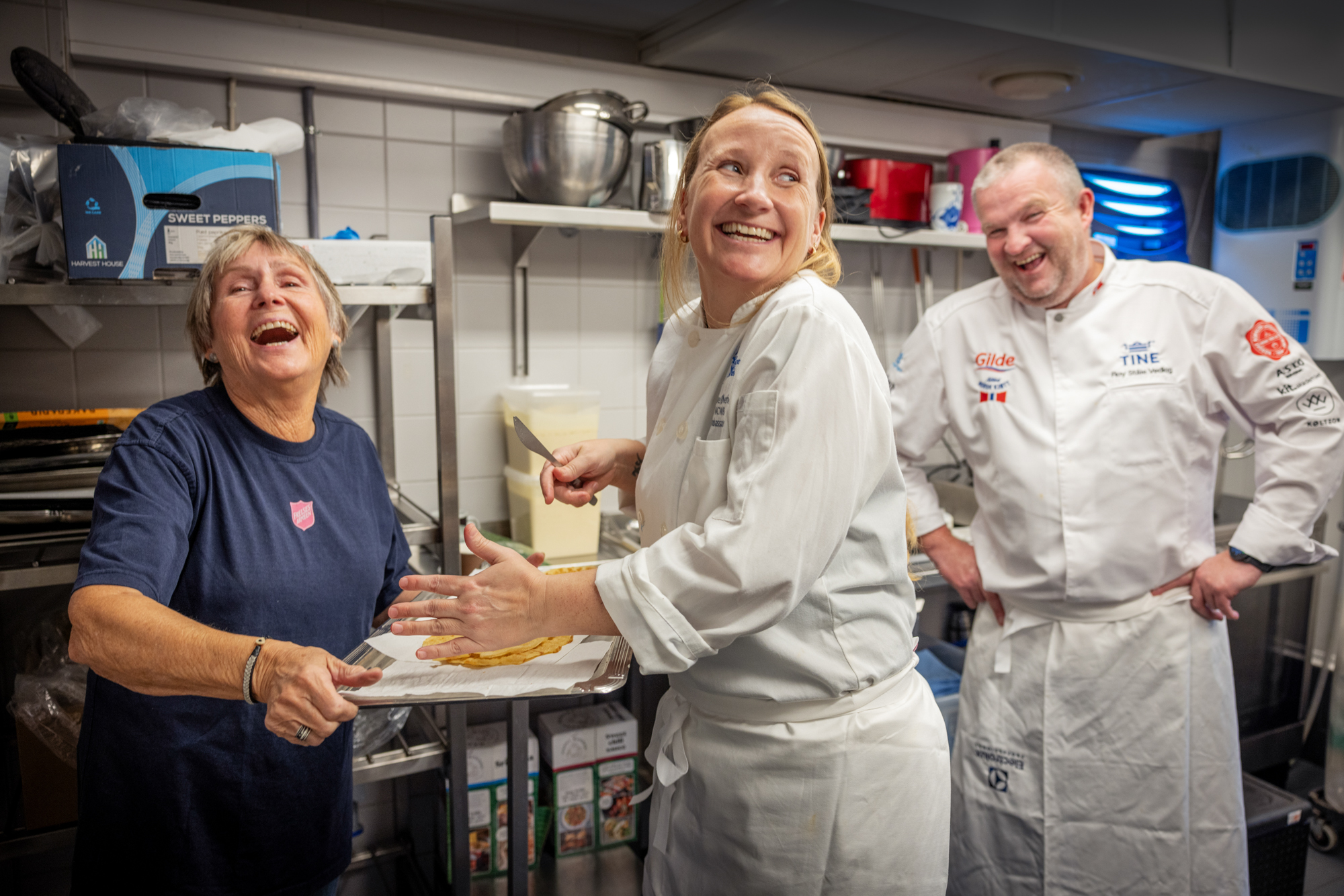 En frivillig i T-skjorte fra Frelsesarmeen sammen med de to kokkene. Alle ler over noen vafler på et brett.