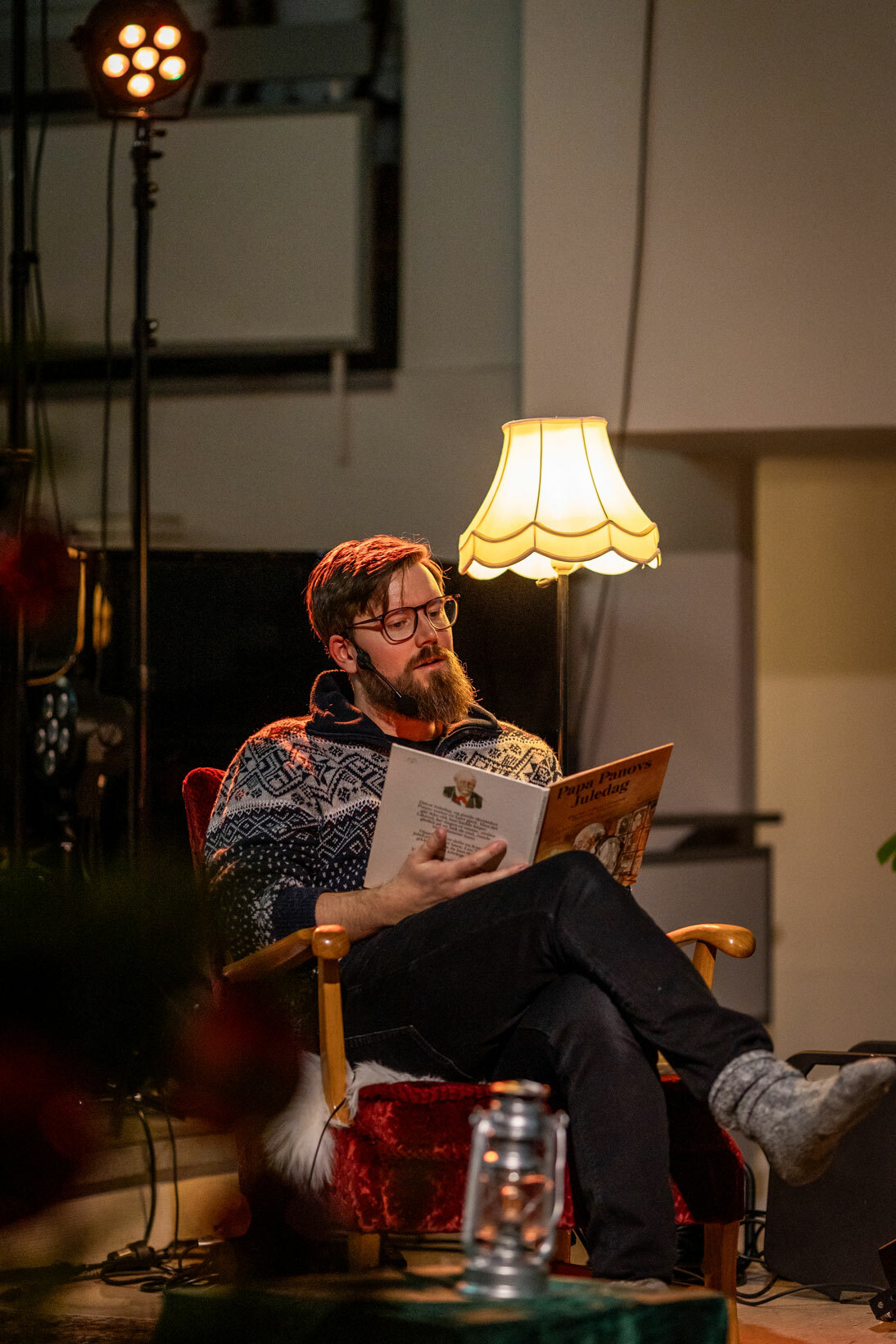 Foto: Mann i 30-årene som sitter i en gammel stol med stålampe bak og leser fra ei bok. Han bruker briller og har på seg en strikkegenser i blått, hvitt og rødt.