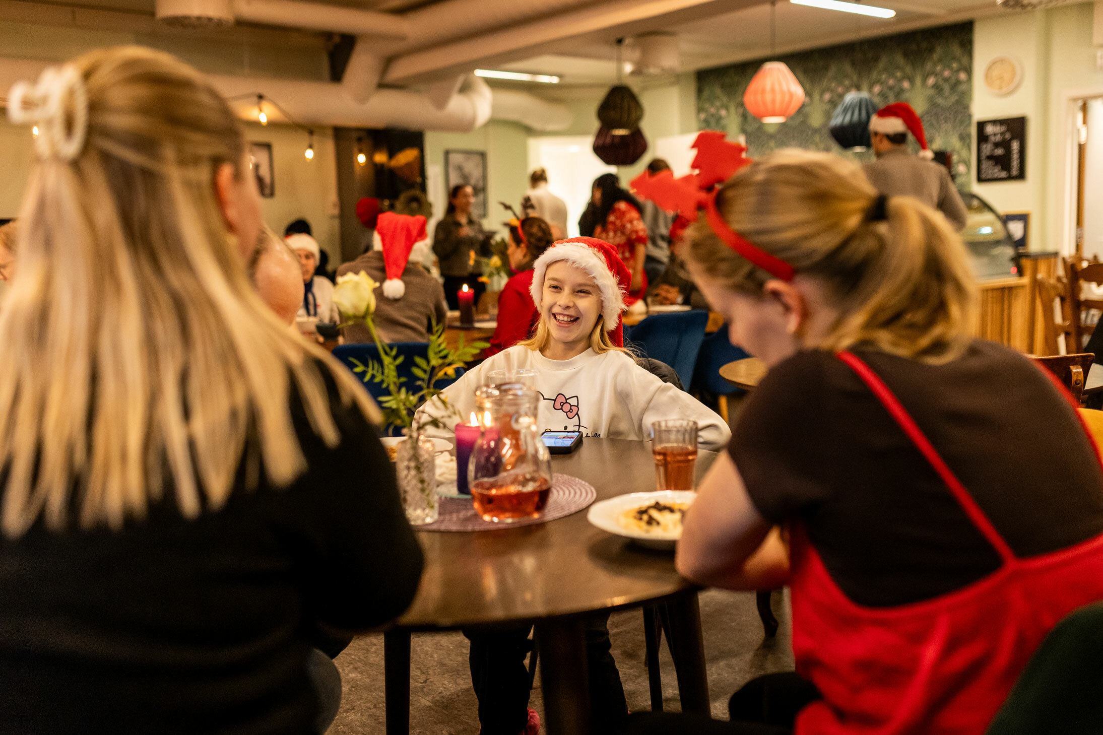 Foto: Ung jente ed nisselue sitter på et bord og snakker med to eldre jenter med ryggen til. Lokalet er fullt av folk.
