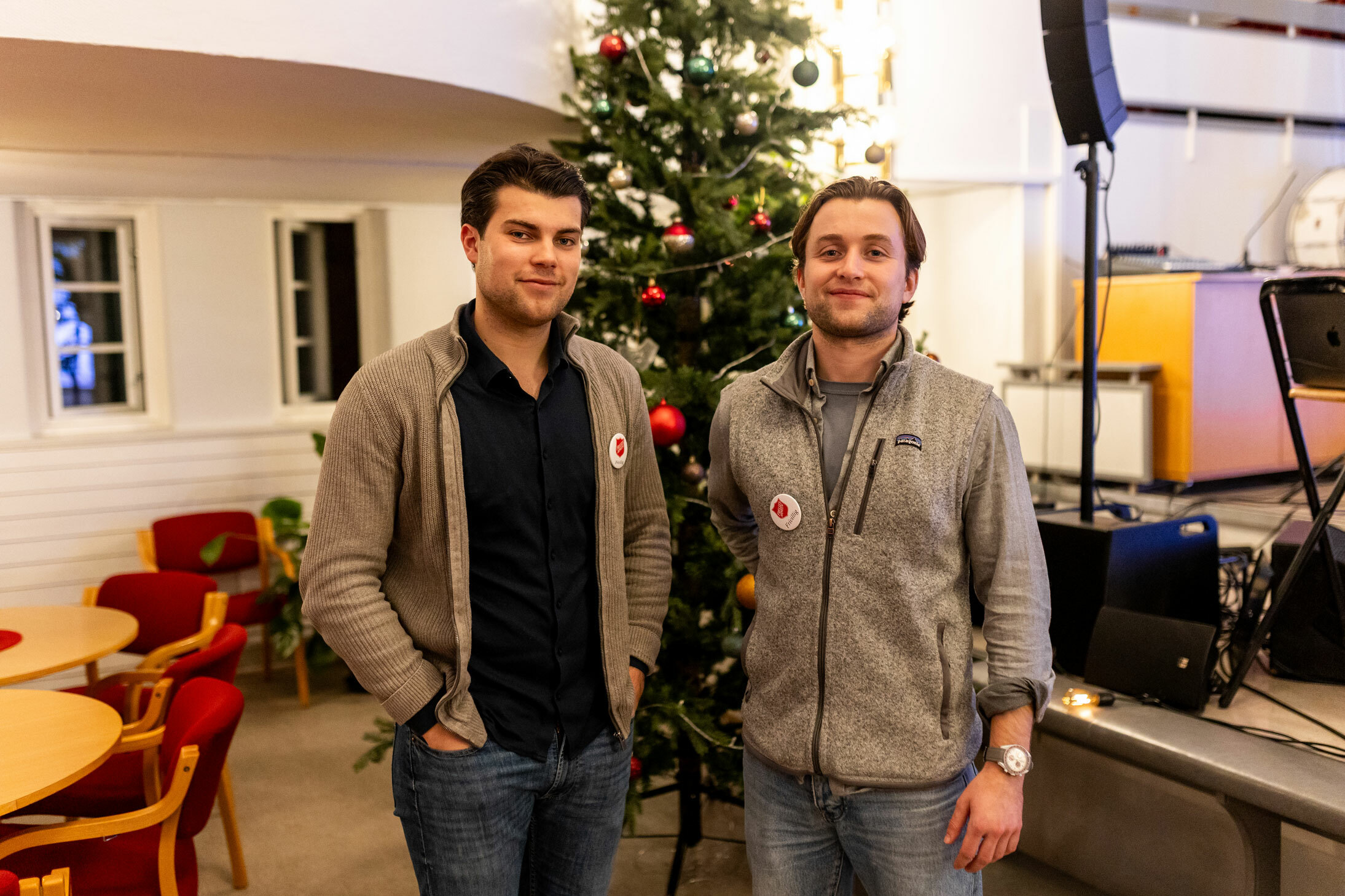 Foto: To menn i midten av 20-årene står foran et pyntet juletre, noen stoler og en scene med høyttalere. Megge har på seg buttons der det står Frelsesarmeen.