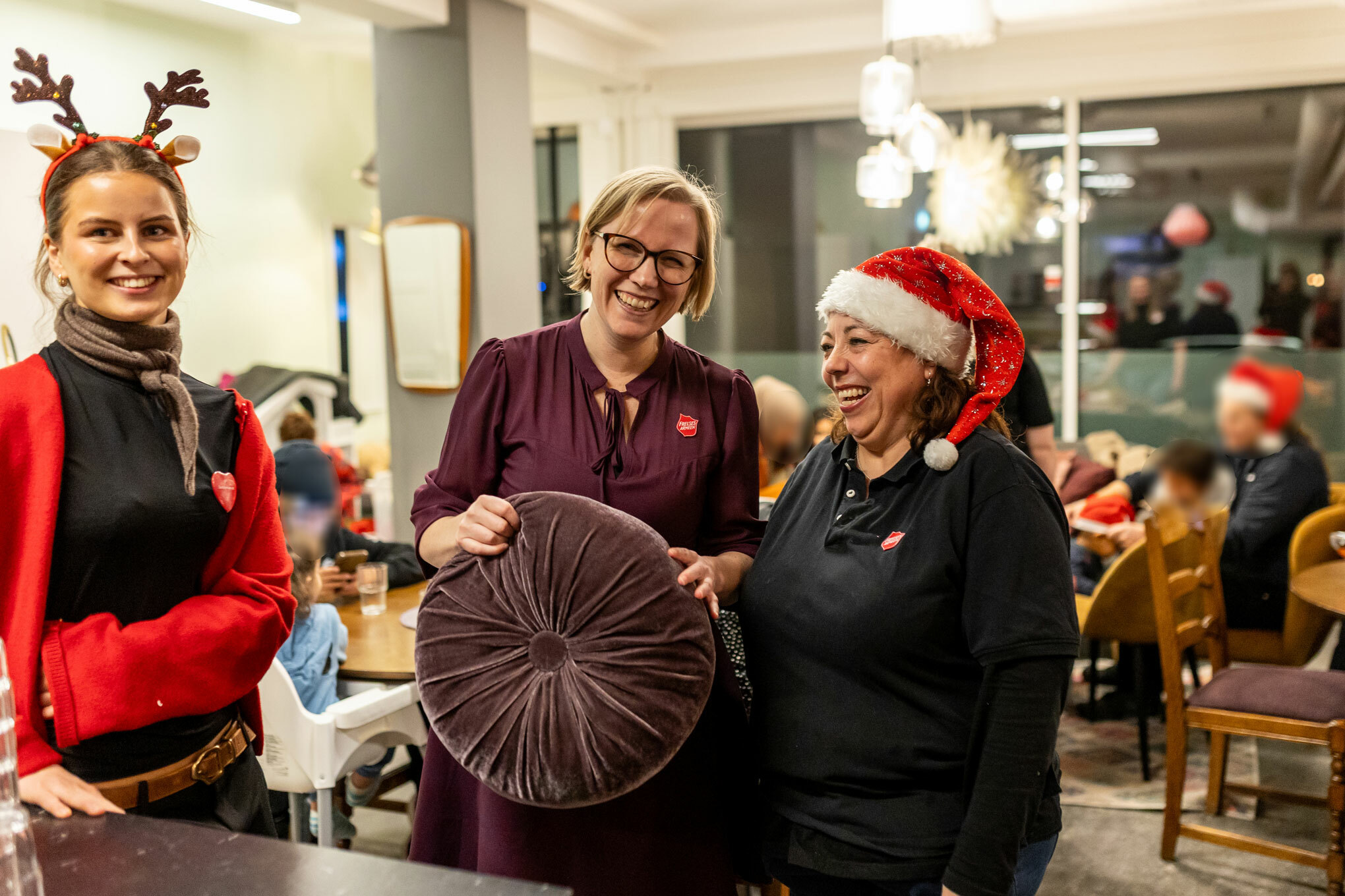 Foto: Tre smilende kvinner står på rad, hun til venstre har en hårbøyle med reinsdyrhorn. De andre to holder på en rund sofapute, der den ytterste har på nisselue..