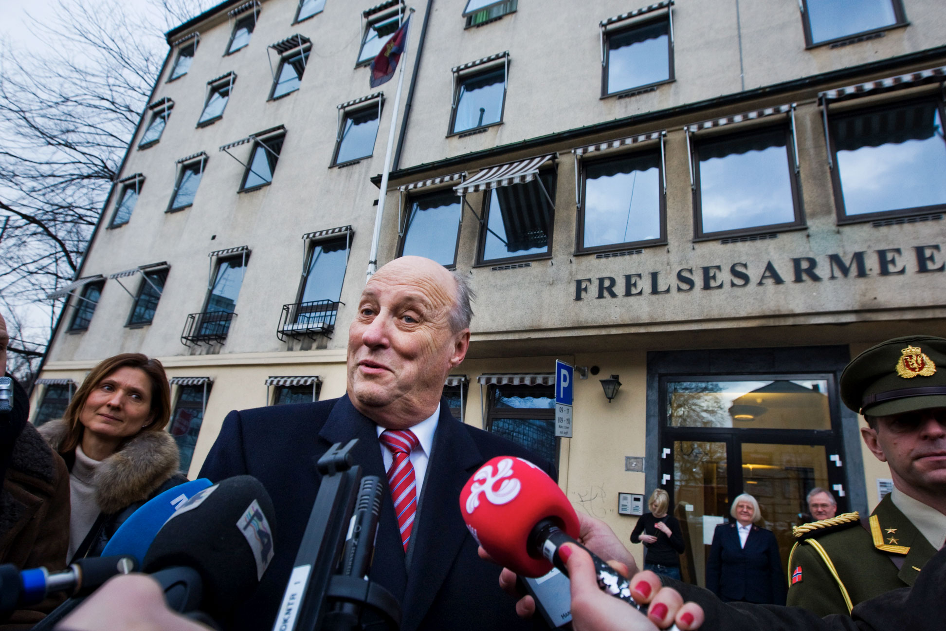 Foto: En mann i 70-årene i dress og slips blir intervjuet av en rekke nyhetskanaler (vi ser mikrofonene) like utenfor et beige murbygg det står Frelsesarmeen på med store bokstaver.