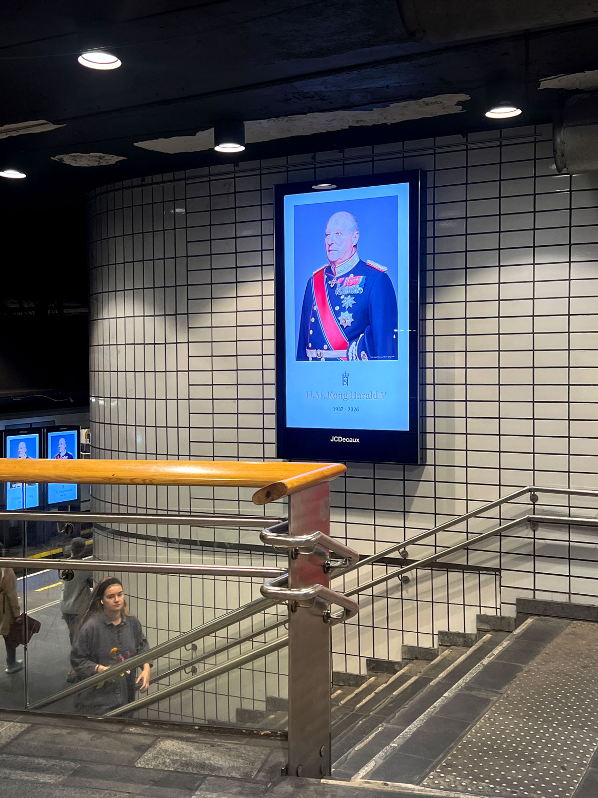 Foto: Tatt ved trappen ned til perrongen inne på en T-banestasjon med hvite fliser på veggen. I en stor, digital "ramme" henger et bilde av kongen i uniform.