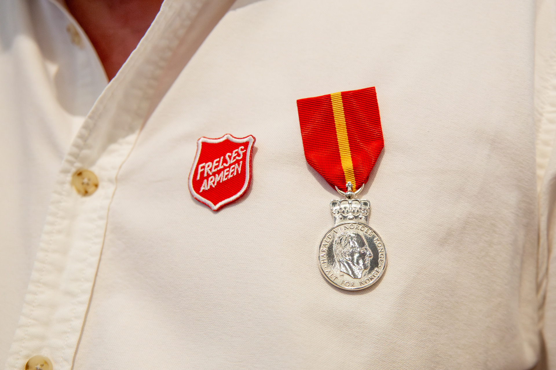 Foto: Nærbilde av en hvit skjorte med logo fra Frelsesarmeen sammen med en medalje i rødt og gult stoff. På enden henger en "mynt" i sølv med kong Haralds ansikt.