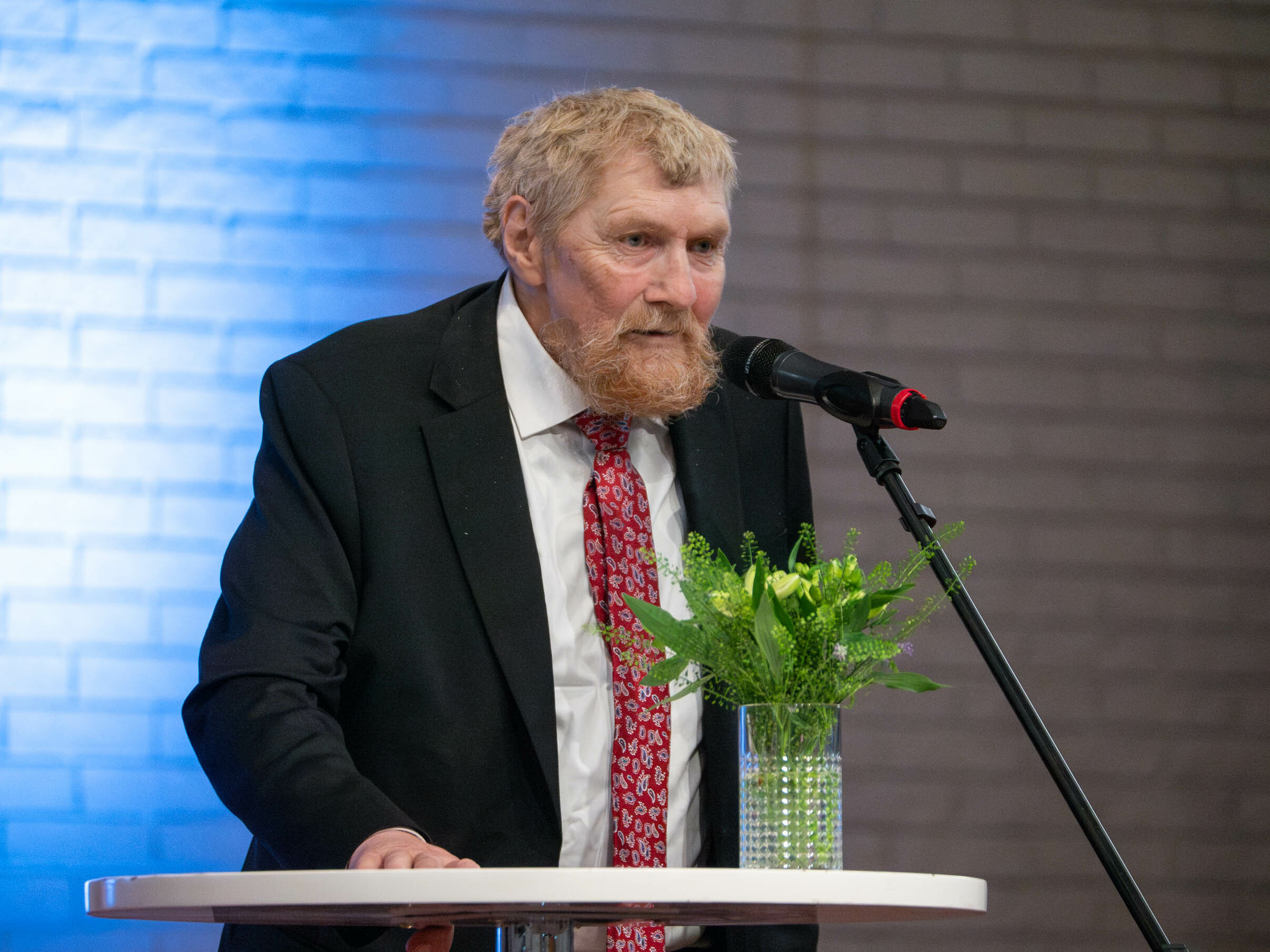 Foto: Eldre herre med rødlig hår og skjegg står på talerstolen. Han har på seg hvit skjorte, rødt slips og mørk dressjakke.