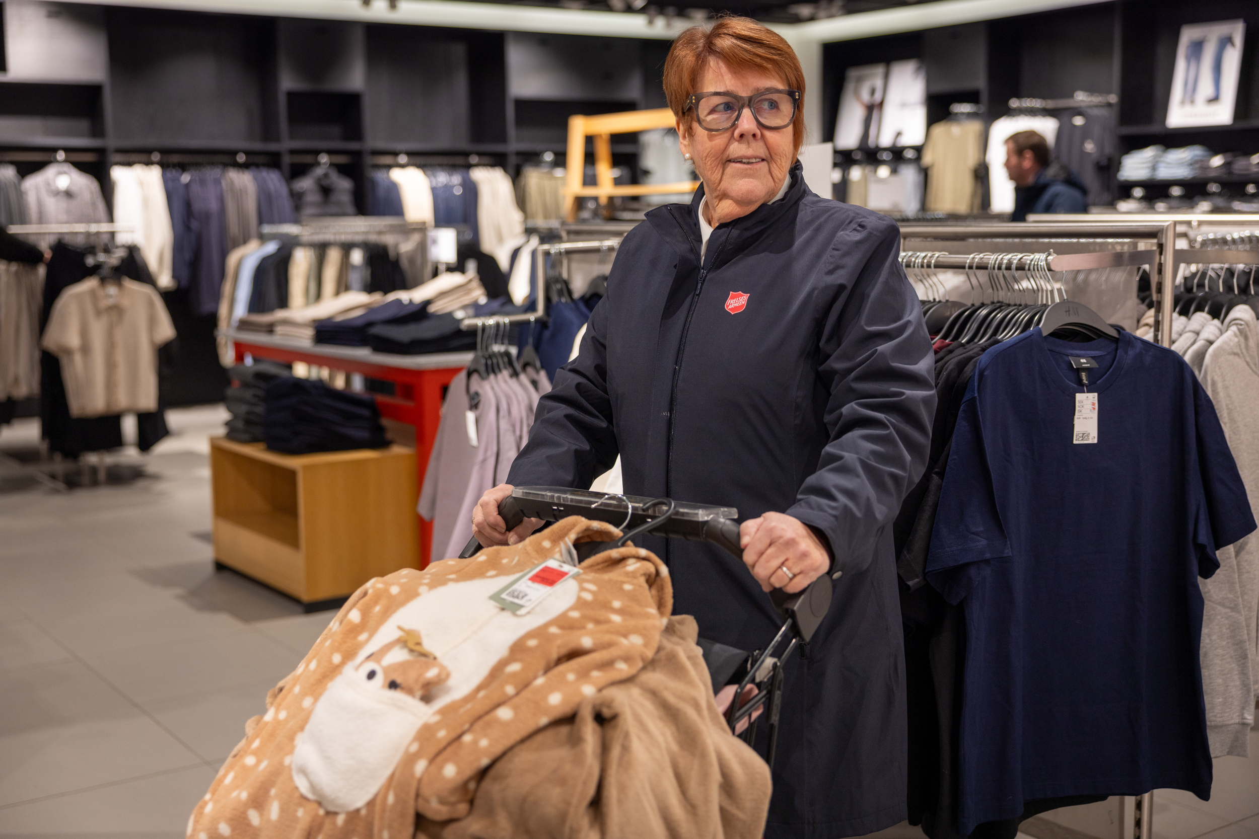 Foto: Eldre kvine med briller og kort hår står i en blå ytterjakke med rød logo fra Frelsesarmeen. Hun holder i en handlekorg med en haug av klær på. Øverst ligger en kosedrakt i beige og hvit.
