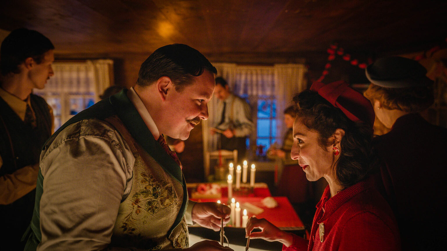 Stor mann med snurrebart og liten sped kvinne ser hverandre inn i øynene. Mørkt rom fullt av folk og stearinlys.