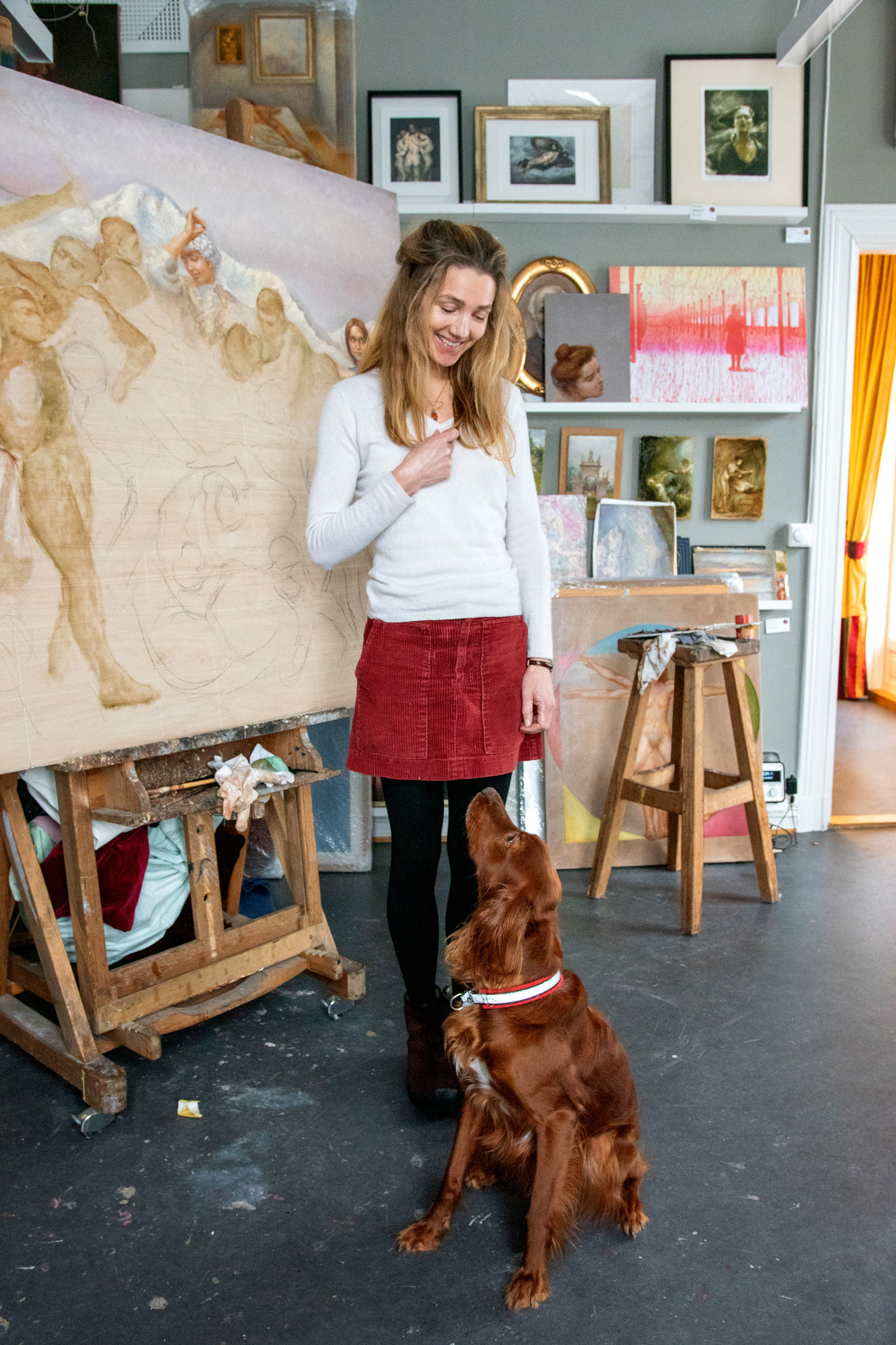 Foto: Dame med langt, mellomblondt hår står foran et maleri og ser ned på en gyllenbrun, stor hund med halsbånd. Den sitter lydig og ser opp på henne. De er i et atelier og hun har på seg hvit genser, rødt skjørt og svart, tykk strømpebukse.