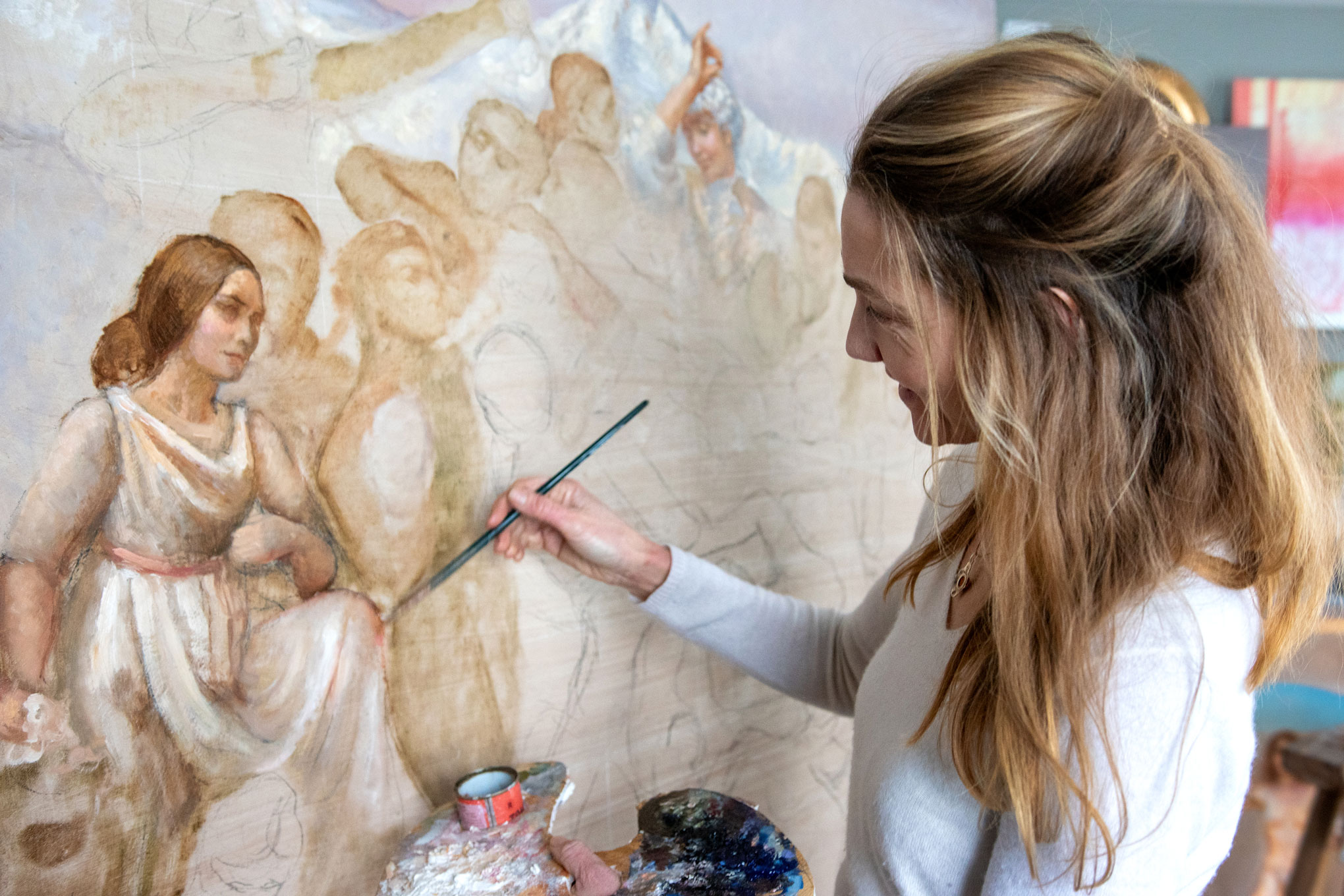 Foto: Kvinne med blondt, langt hår maler med pensel på et bilde i duse farger. Frem fra lerretet står en tydelig kvinneskikkelse fram. De andre menneskene på bildet er skisset inn og skal males tydeligere.