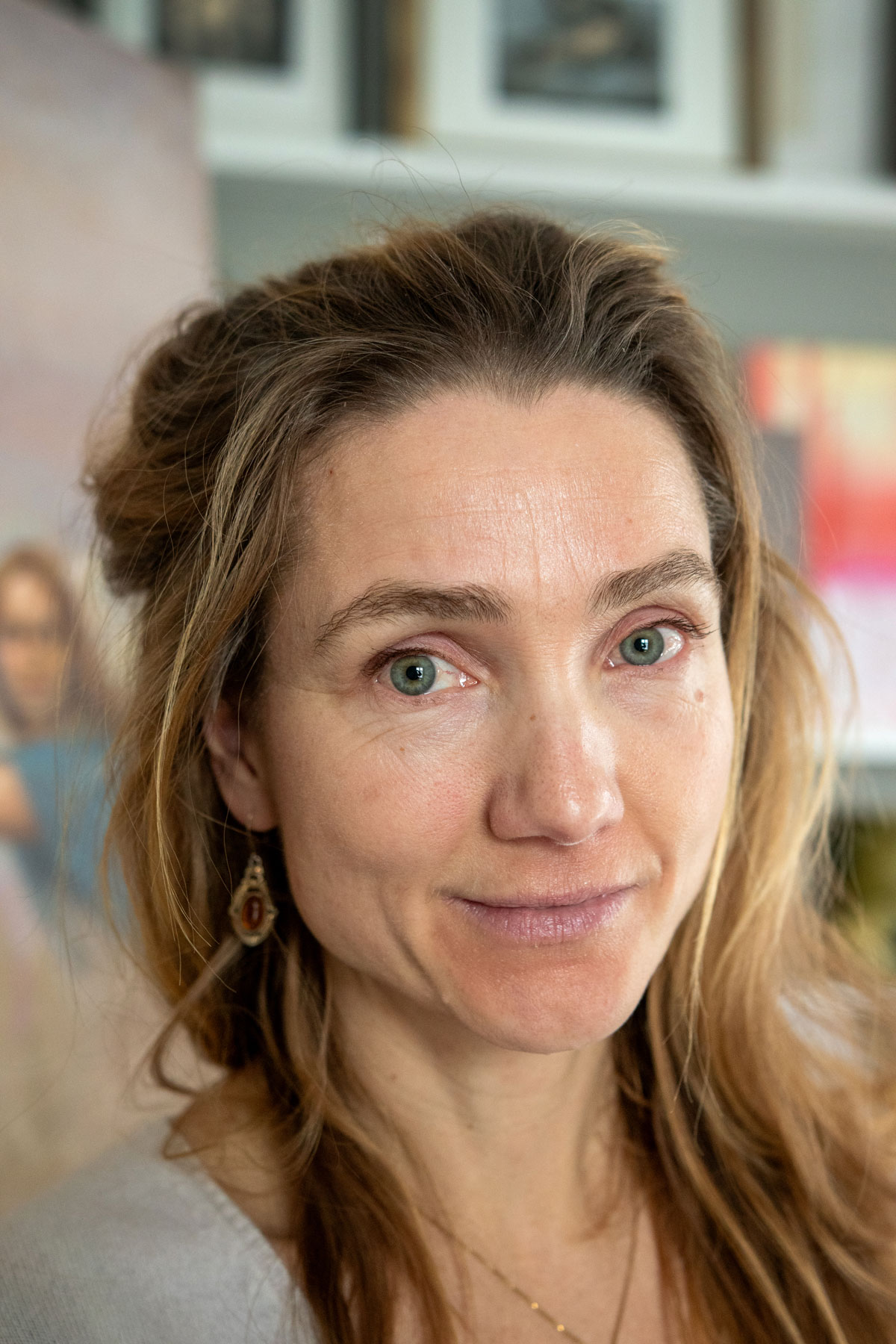 Foto: Nærbilde av en mellomblond kvinne med blå øyne, mørke, markerte øyenbryn og lysere striper i håret. Hun er i et atelier og ser rett i kamera.