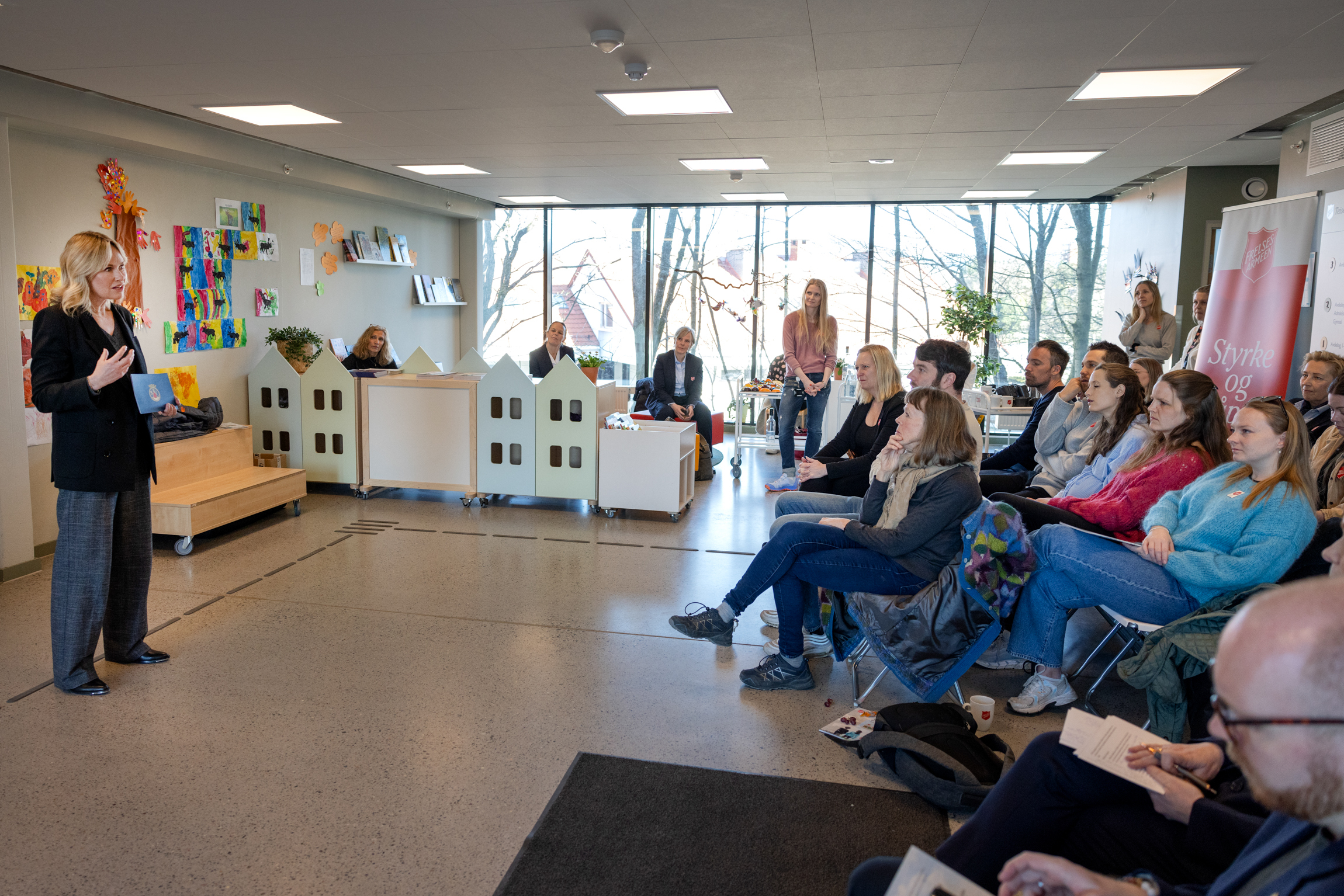 Dame står foran en større forsamling som sitter inne i en stor barnehageavdeling.
