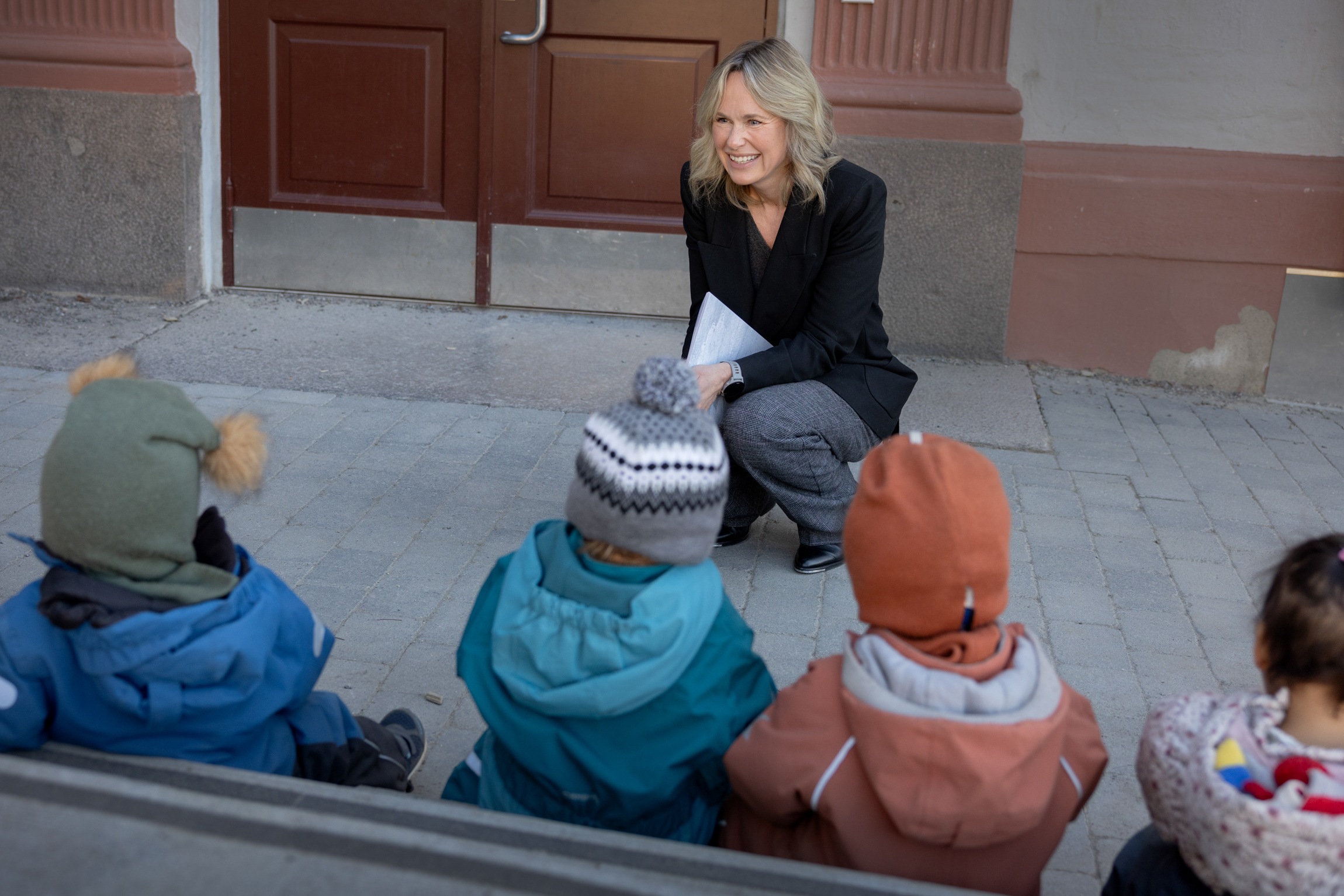 Foto: Kvinne i dressjakke sitter på huk foran fire barn med luer og parkdress som sitter på en benk. Kan ikke se ansiktet deres.