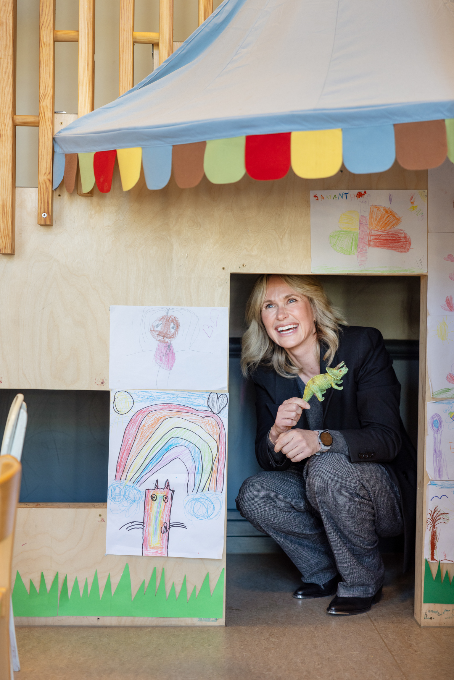 Foto: Voksen kvinne med blondt hår og mørk dressjakke sitter på huk inne i et lite lekerom for barn.