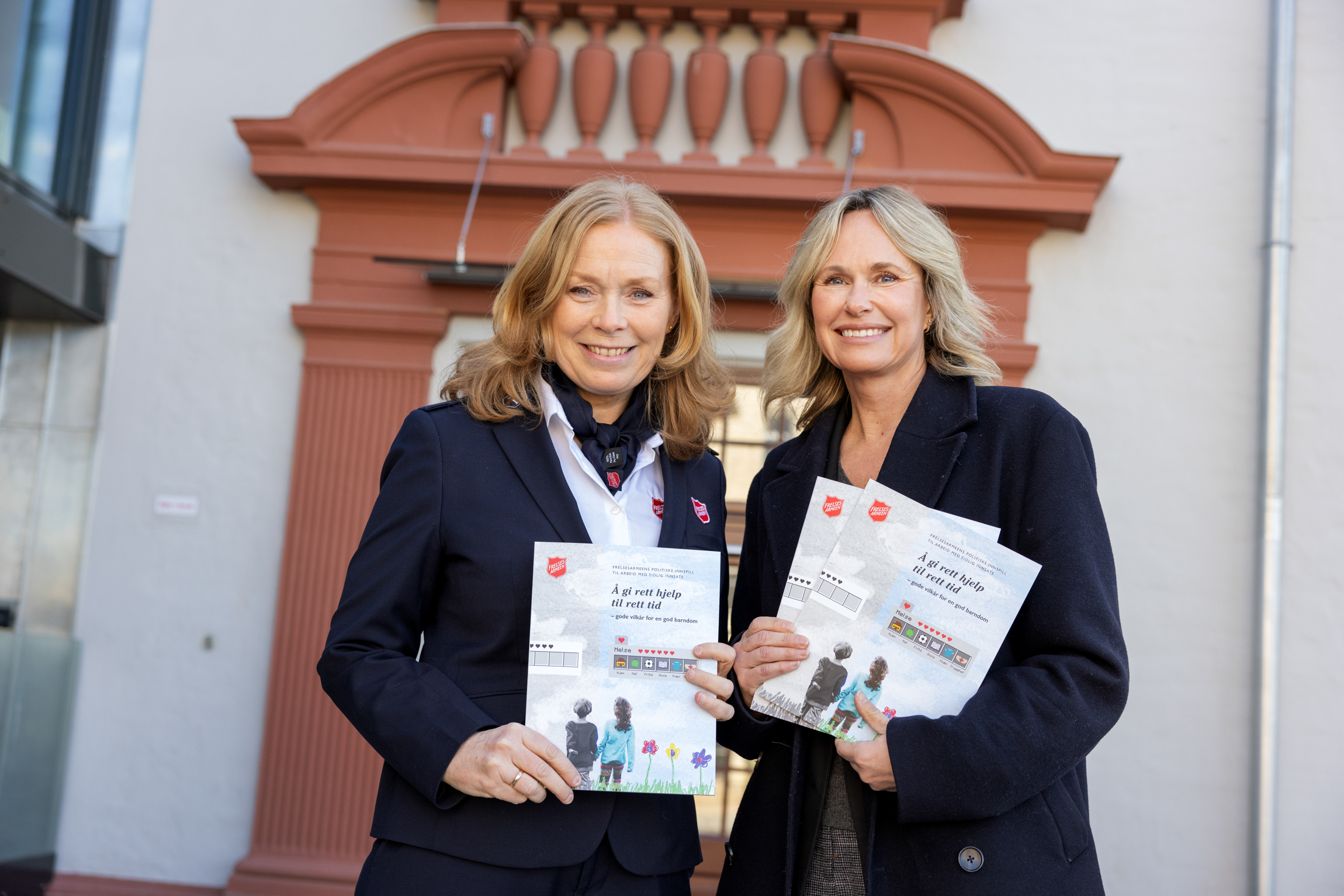 Foto: To smilende kvinner i hver sin mørke dressjakke smiler til fotografen. De står utenfor en gammel murbygning og holder til sammen fram tre rapporter med Frelsesarmeens logo oppe i venstre hjørne.