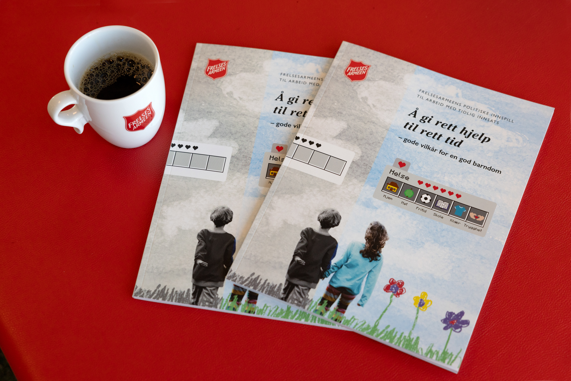 Foto: Hvit kaffekopp med Frelsesarmeens logo står oppå en rød bordflate. De ligger også to utgaver av en rapport med bilde av to barn som står med ryggen til. Tittel på rapporten er «Å gi rett hjelp til rett tid – gode vilkår for en god barndom».