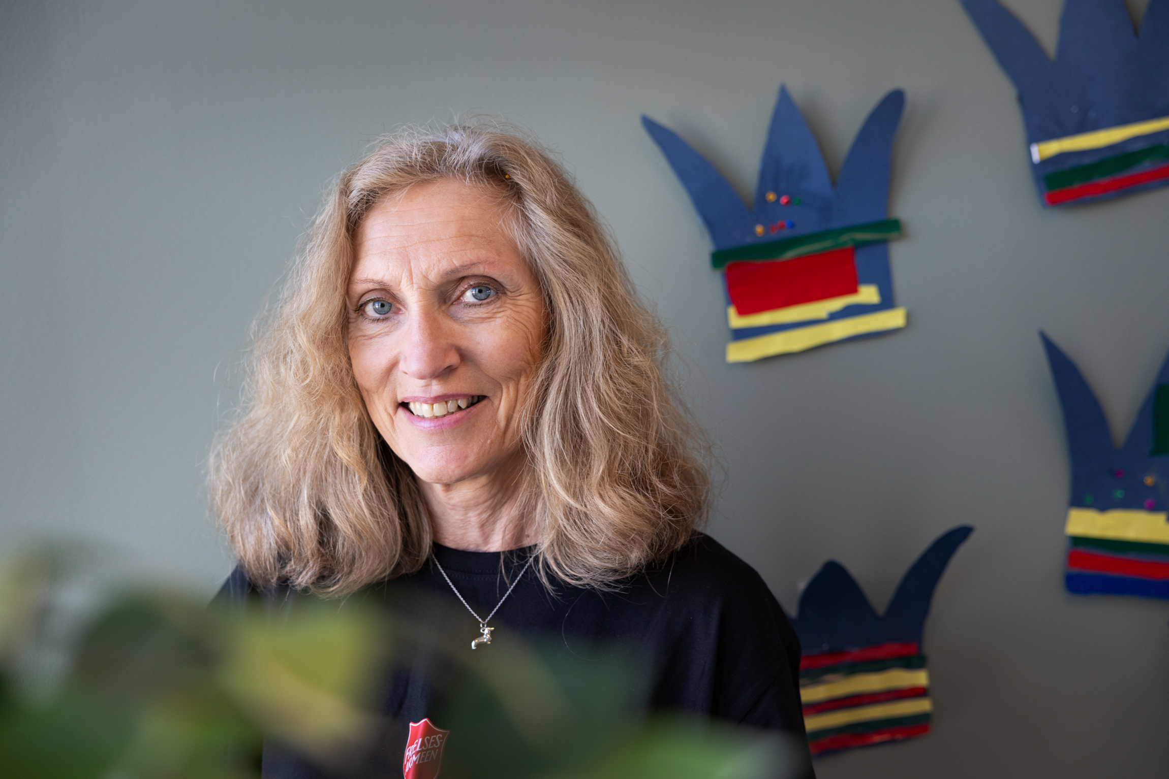 Foto: Moden kvinne med krøllete, gråblondt hår sitter foran en vegg med en rekke kroner på veggen. Disse har barn malt i blått, rødt og gult og klippet ut av papir.