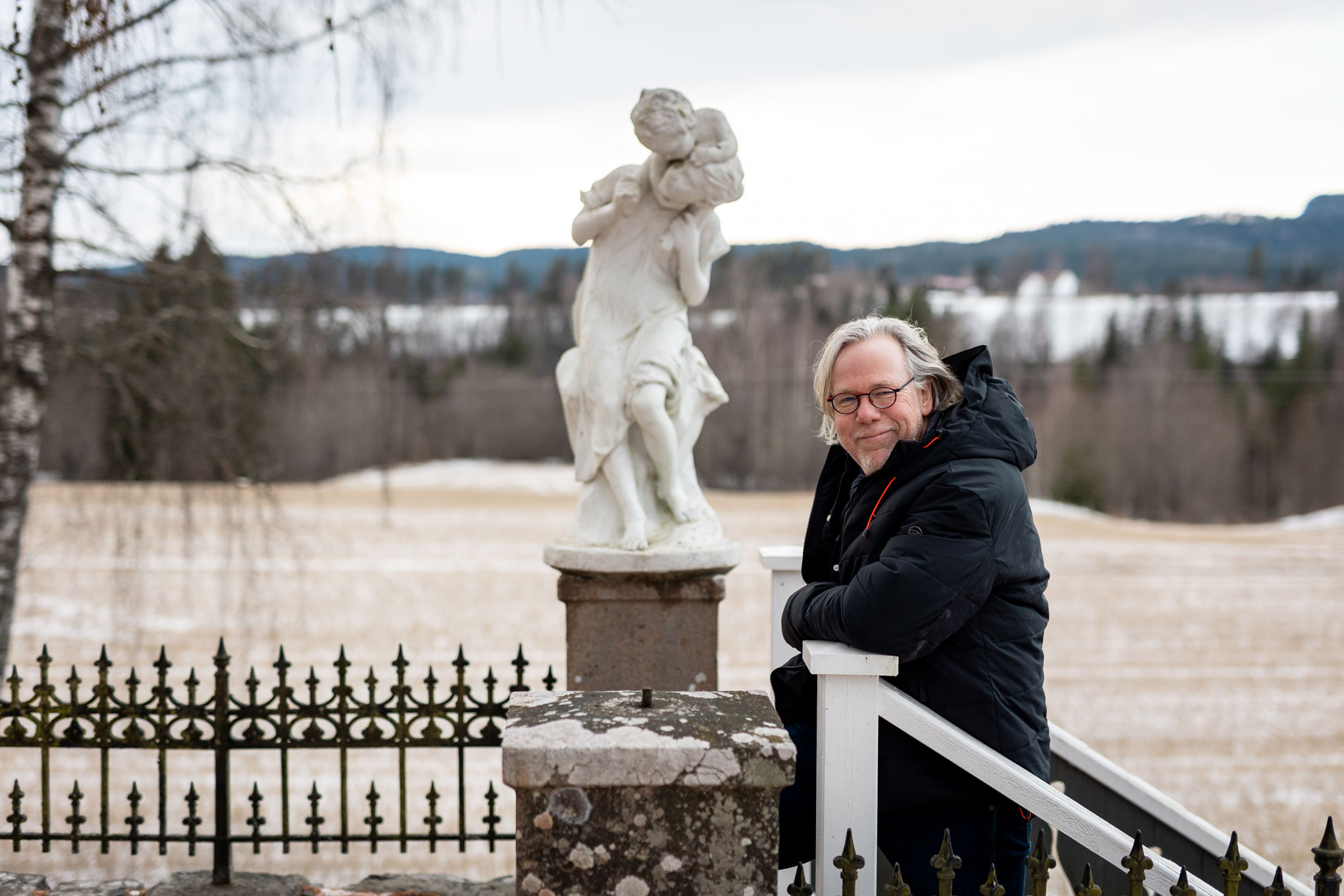 Foto: En mann med grått hår, briller og svart vinterjakke står i en trapp utendørs og lener seg mot gjerdestolpen. Ved siden av ham er en hvit statue av en kvinne som har et lite barn på skuldrene. Kvinnen har hodet til siden og ser på barnet.
