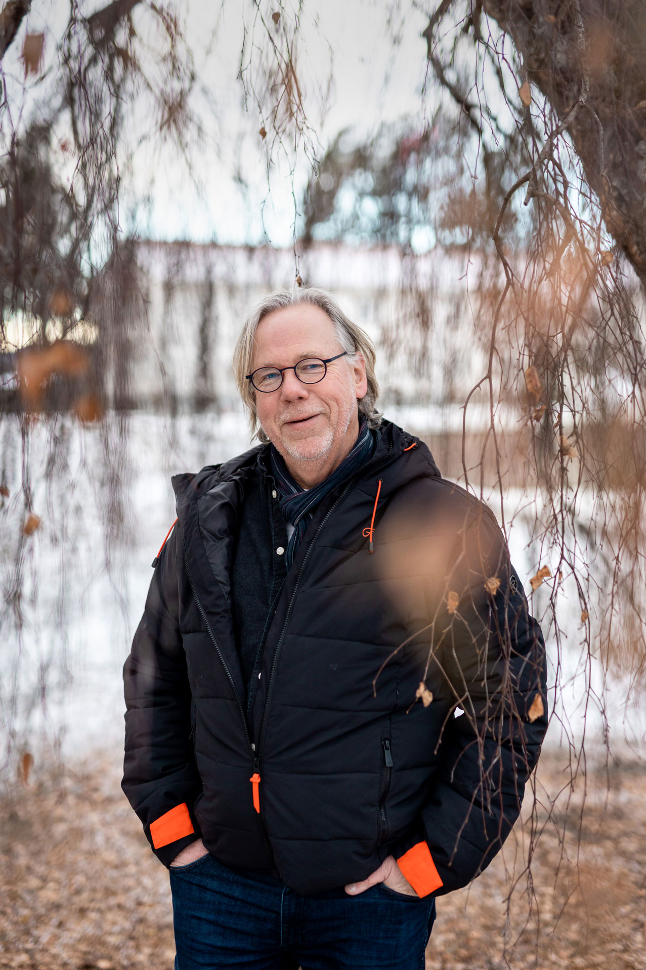 Foto: En mann med grått hår og svarte klær står mellom grenene til en hengebjørk om vinteren. Han har hendene i lommene og smiler mot kamera. Bak ham sees et stort, hvitt bygg som er uskarpt i bildet.