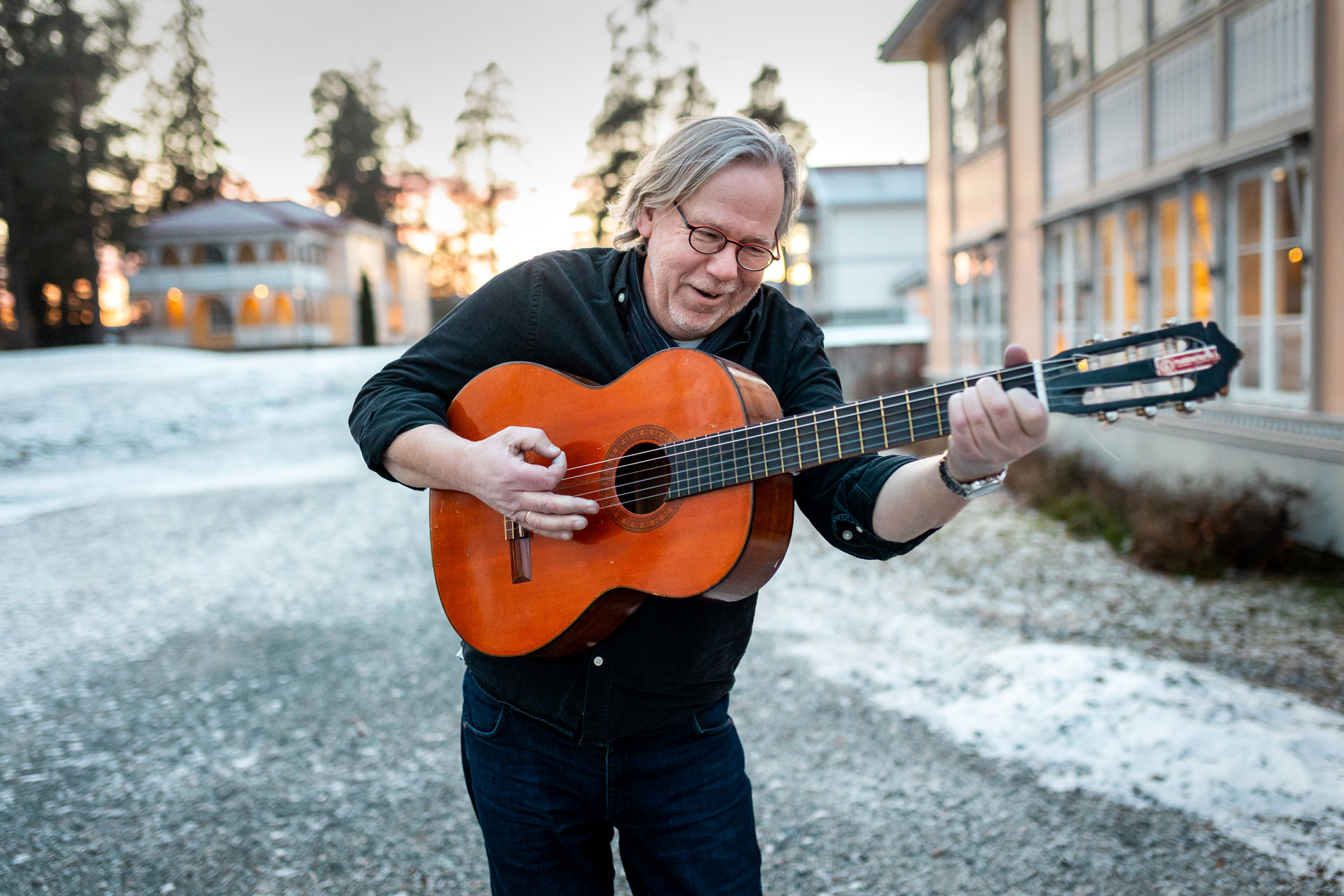 Foto: En mann med grått hår og svarte klær, står utendørs og holder en gitar. Han ser litt ned, og ser ut som han spiller og synger. Bak ham sees bakken med litt snø og flere bygninger.