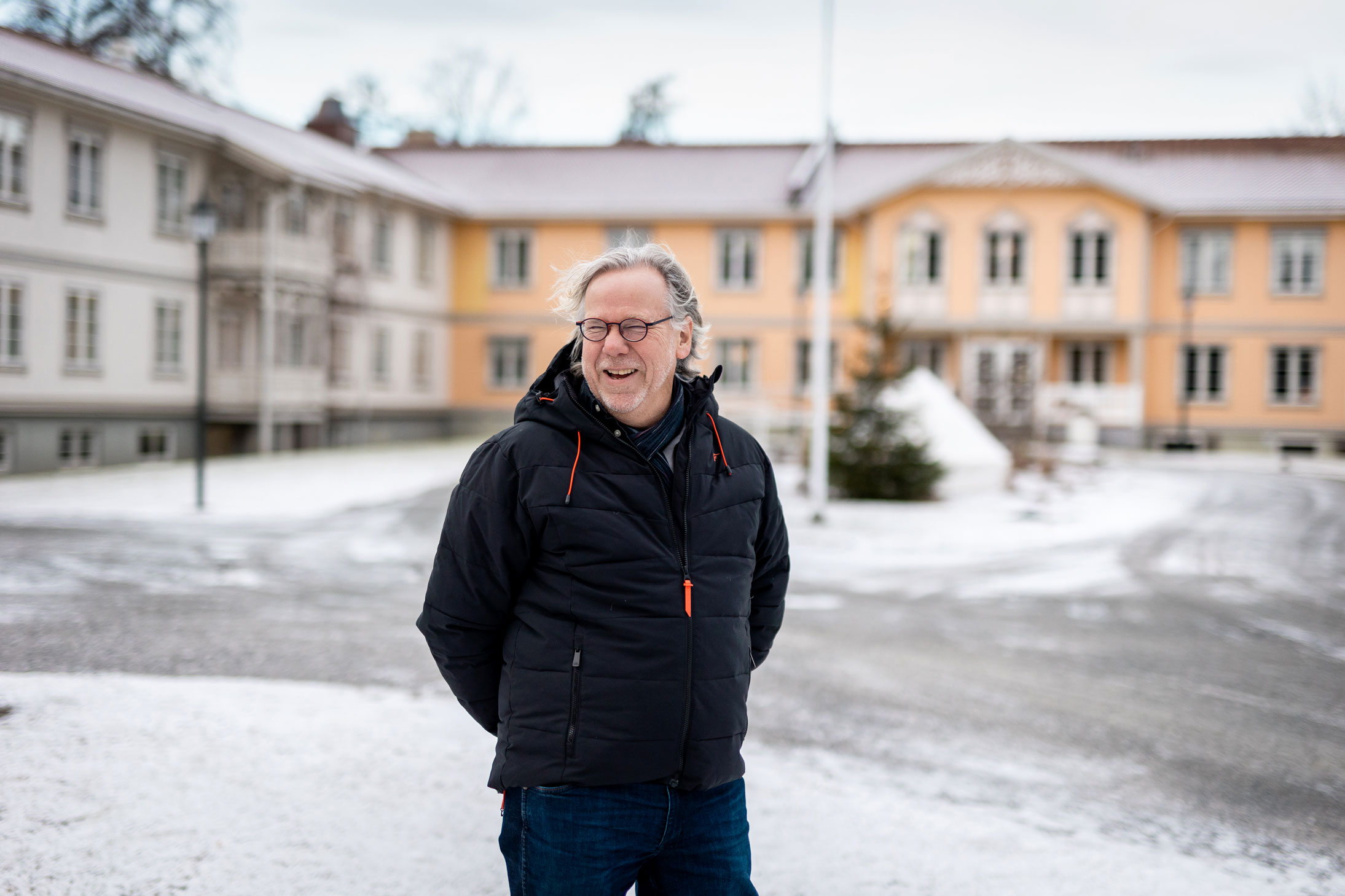 Foto: En i 50-årene med litt lengre, grått hår og briller står i en svart boblejakke med oransje detaljer foran et par store, gamle bygninger. Den ene er malt i gult og den andre er malt i hvitt. Han ler godt så tennene viser.
