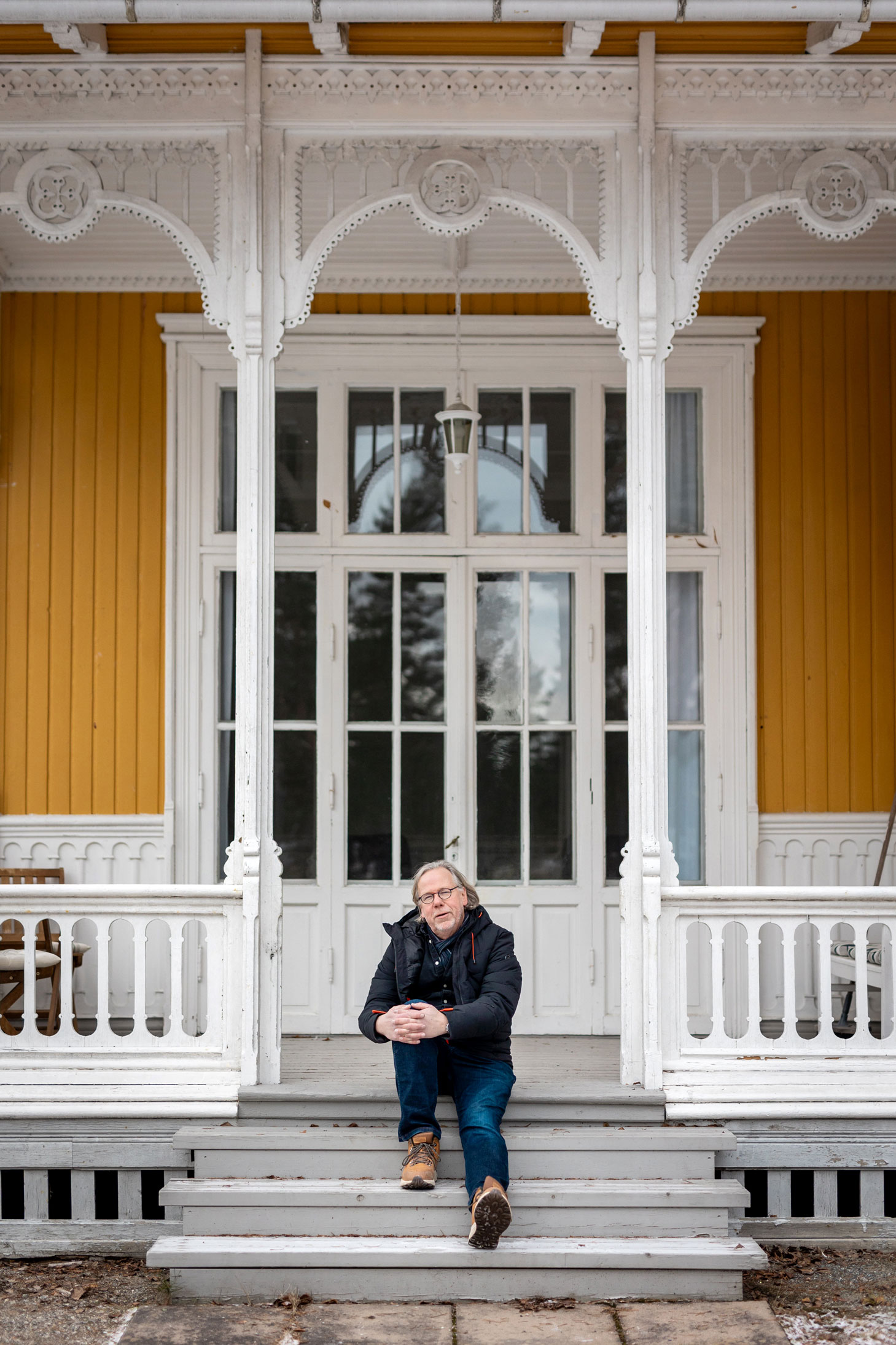 Foto: En mann sitter på en trapp utenfor et gammelt gult og hvitt trebygg med hvite utskjæringer. Bak ham er store glassdører inn til huset.
