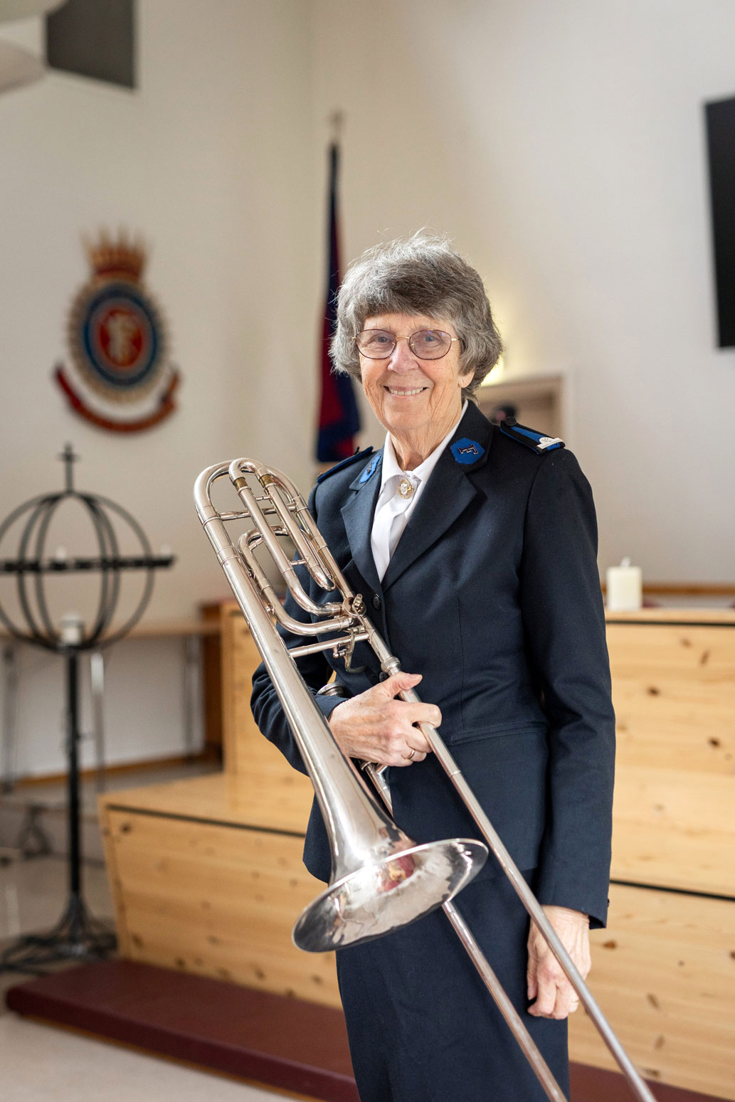 Foto: Kvinne i uniform fra Frelsesarmeen står med en trombone i hendene inne i et kirkerom som tilhører Frelsesarmeen. Det ser en på flagget som står i det ene hjørnet og kronen på veggen der det står "Blod og ild".