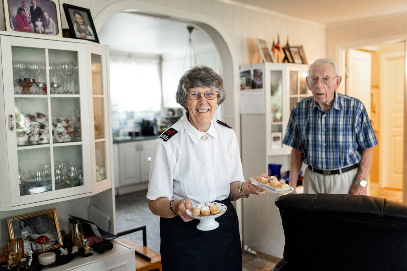 Foto: Kvinne i slutten av 70-årene med briller og uniform fra Frelsesarmeen bærer to fat med kaker med bakst i en stue. Rett bak henne går en jevngammel mann med grått hår, briller og blårutete skjorte.