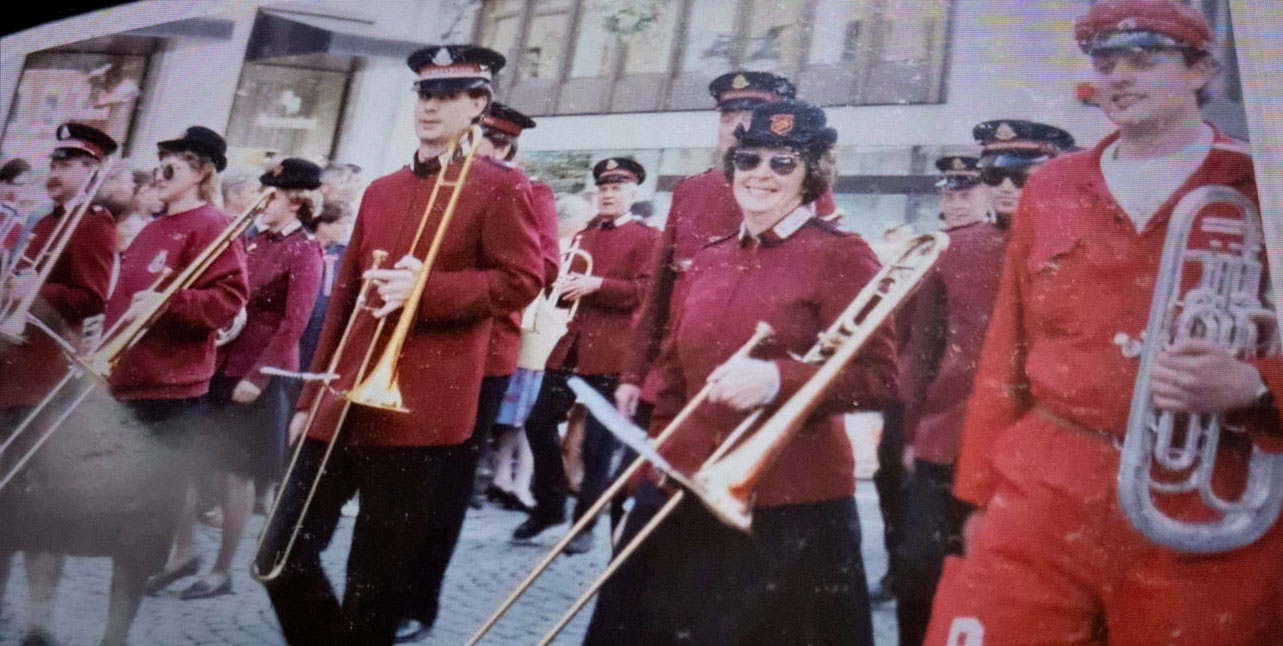Foto: Gammelt fargebilde av musikanter i burgunder jakker og mørkeblå bukser og skjørt står går i en gate.