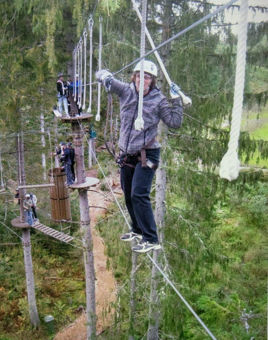 Foto: Kvinne høyt oppe i årene står høyt oppe på en line spent mellom noen grantrær. Hun holder seg fast i en line over og flere tau.