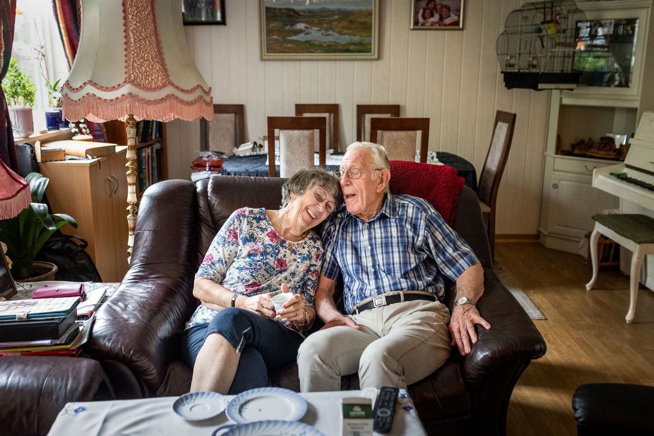 Foto: Kvinne og mann i 70–80-årene sitter i en burgunder skinnsofa. På veggen bak i stua er det panel malt i offwhite. Det står et fuglebur i et hjørne og ellers er det bilder på veggene.