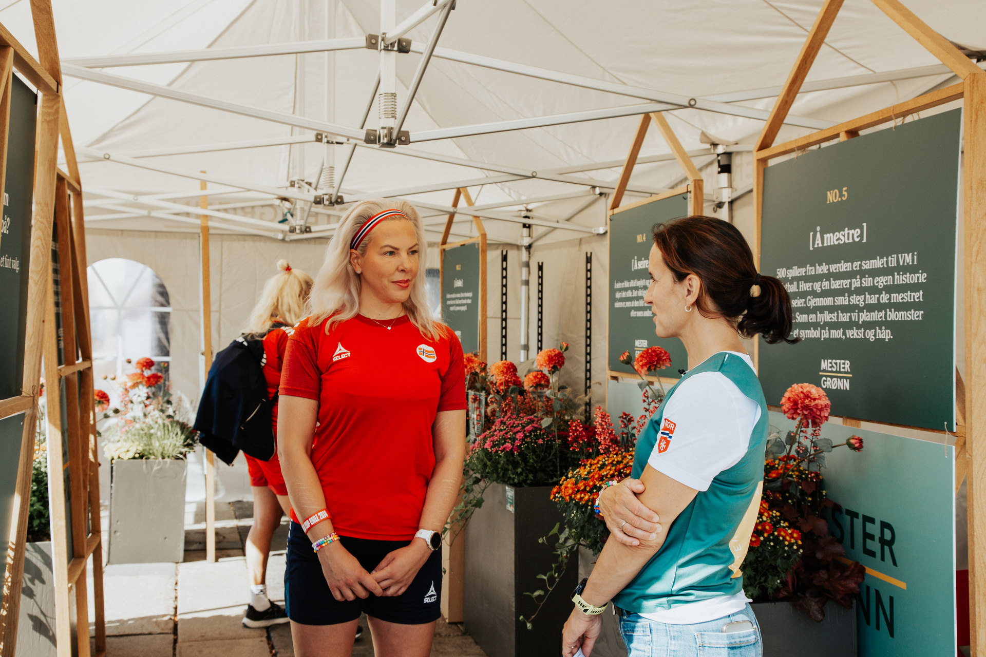 Foto: Cathrine Johansen og Marit Bjørgen står inne i et stort telt og prater. Bjørgen står med siden mot kamera, Johansen står vendt mot henne i rød og svart fotballdrakt.