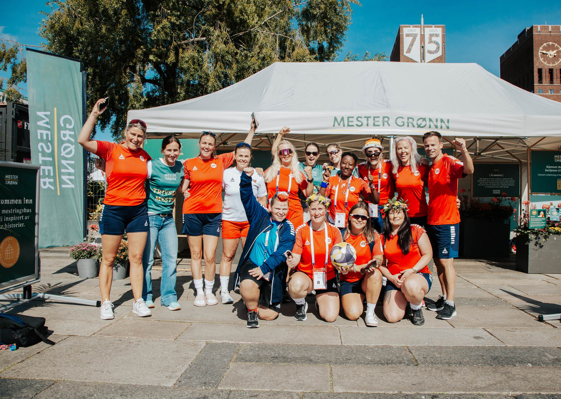Foto: Oppstilt lagbilde av det norske kvinnelandslaget med oransje fotballtrøyer, sammen med Marit Bjørgen og de to langrennsjentene. Marit Bjørgen står som nummer to til venstre sammen med landslagstrener Signe Haukvik Haugen. Fire spillere sitter på huk foran og de andre står bak.