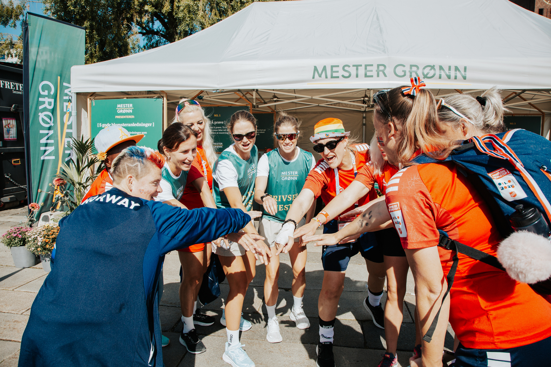 Foto: Marit Bjørgen står til venstre i bildet i en sirkel av gatefotballspillere. Alle lener seg fram og holder en hånd inn mot midten av sirkelen. I bakgrunnen sees bannere og et telt med Mester Grønn-logo